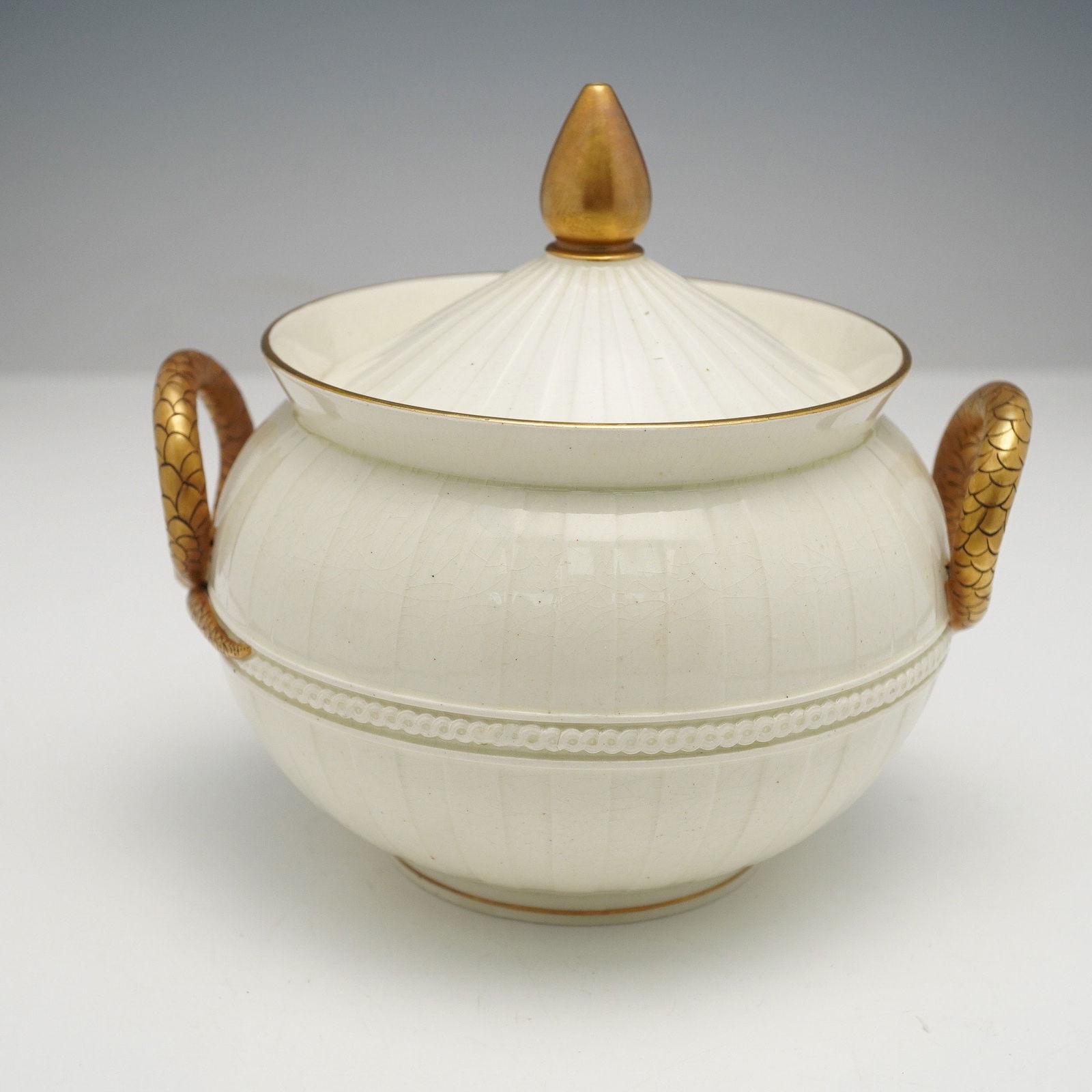 Wedgwood Creamware Sugar Bowl with Serpent Handles Lid: A Wedgwood covered sugar bowl with lid featuring a creamy white ceramic body with vertical fluting, accented by gold trim on the rim and foot. The piece is fitted with a matching fluted domed lid topp