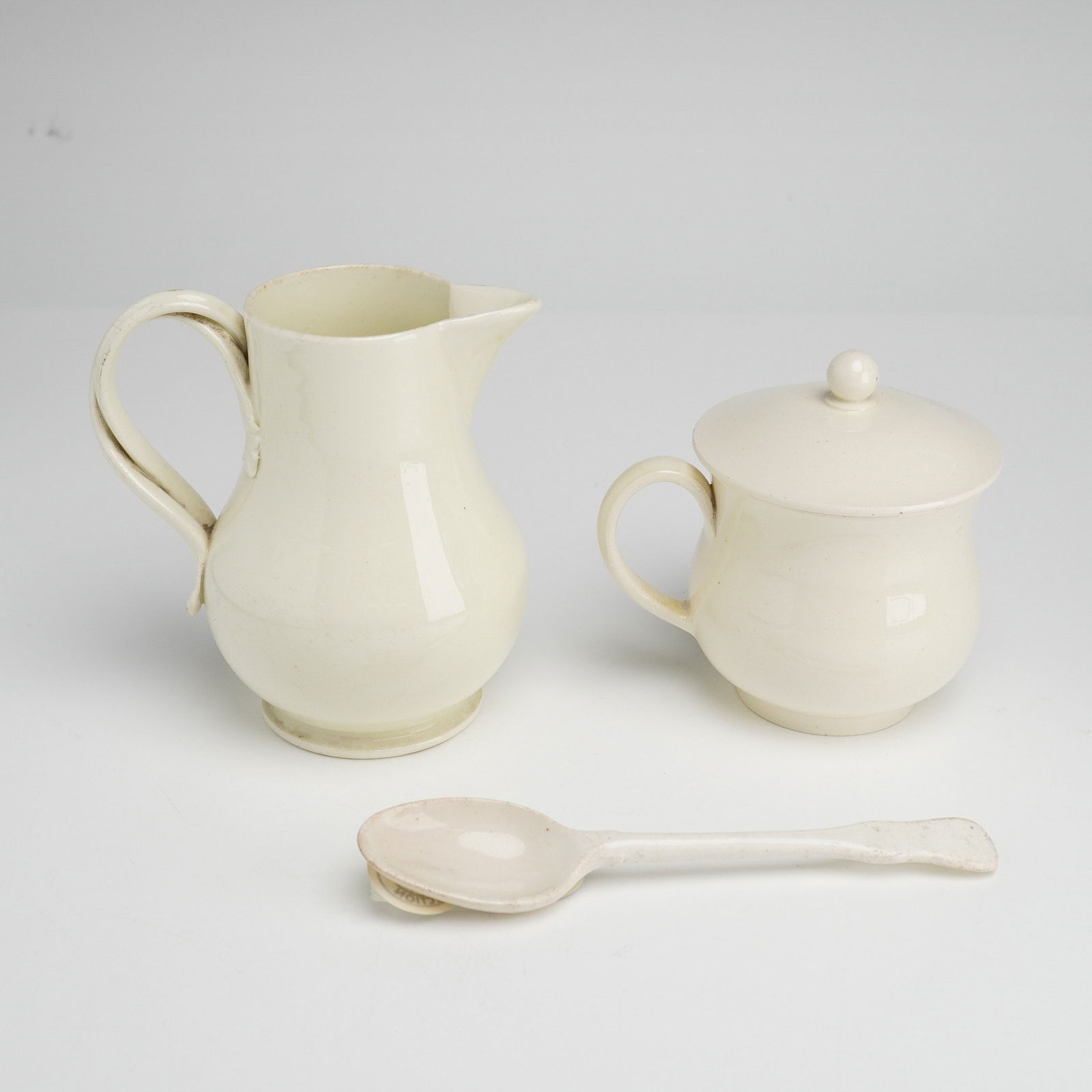 Three Wedgwood Creamware Pots De Creme with Lid, Creamer, and Spoon: A three-piece set of Wedgwood creamware table articles including a pot de creme with lid, a creamer, and a spoon. Each piece is finished in a uniform cream-colored glaze. The creamer features a bulbou