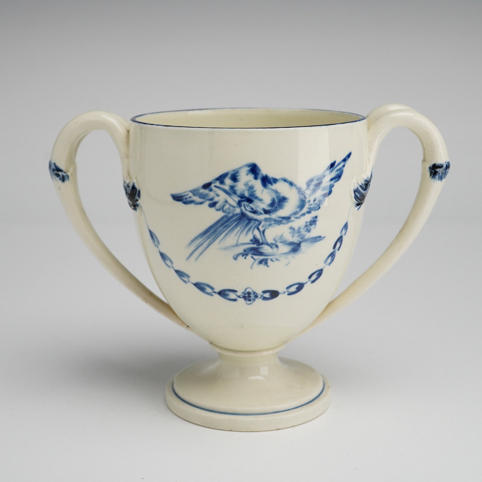 Wedgwood Creamware Bird and Flower Motif Loving Cup: A Wedgwood creamware footed cup featuring dual scroll handles, decorated in a blue-and-white motif. One side showcases a central bird design, while the reverse presents a floral sprig, each framed by