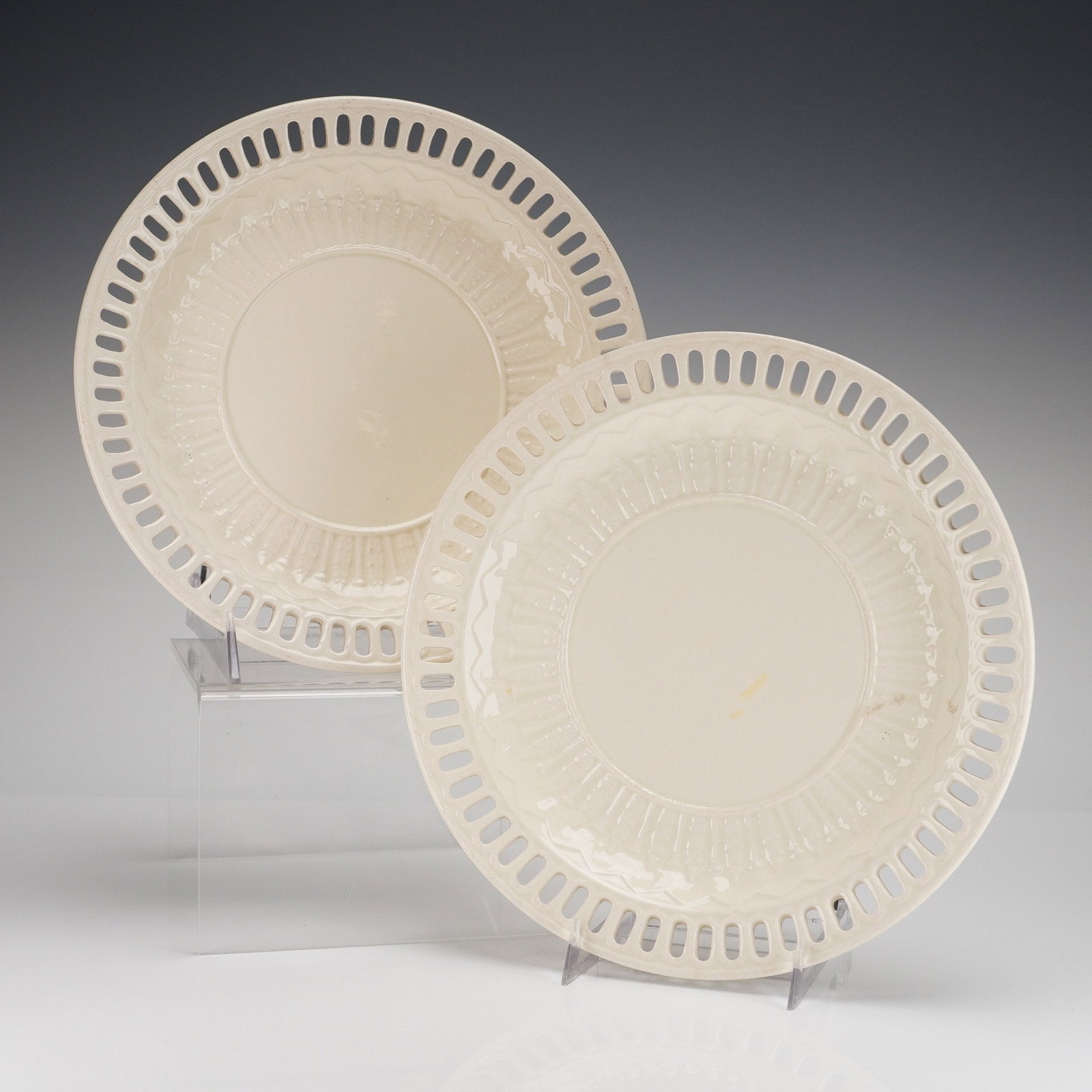 A Pair of 19th Century Wedgwood Creamware Reticulated Plates: A pair of circular creamware plates manufactured by Wedgwood, England. The plates feature a reticulated border composed of elongated oval openings. The undersides bear a printed mark reading WEDGWOOD