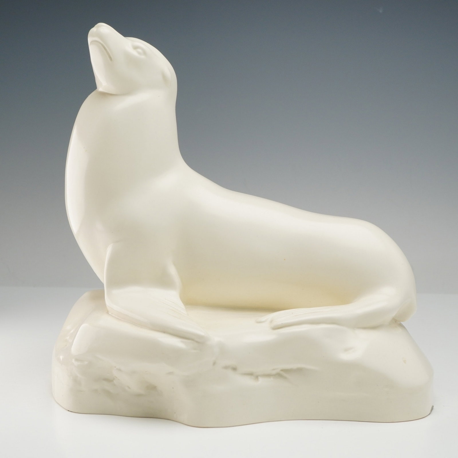 A Wedgwood Glazed Ceramic Sea Lion Figurine by John Skeaping: A creamy white, glazed ceramic figurine depicting a sea lion, designed by John Skeaping for Wedgwood. The marine mammal is shown sitting upright on a textured, rock-like base with its head tilted upwa