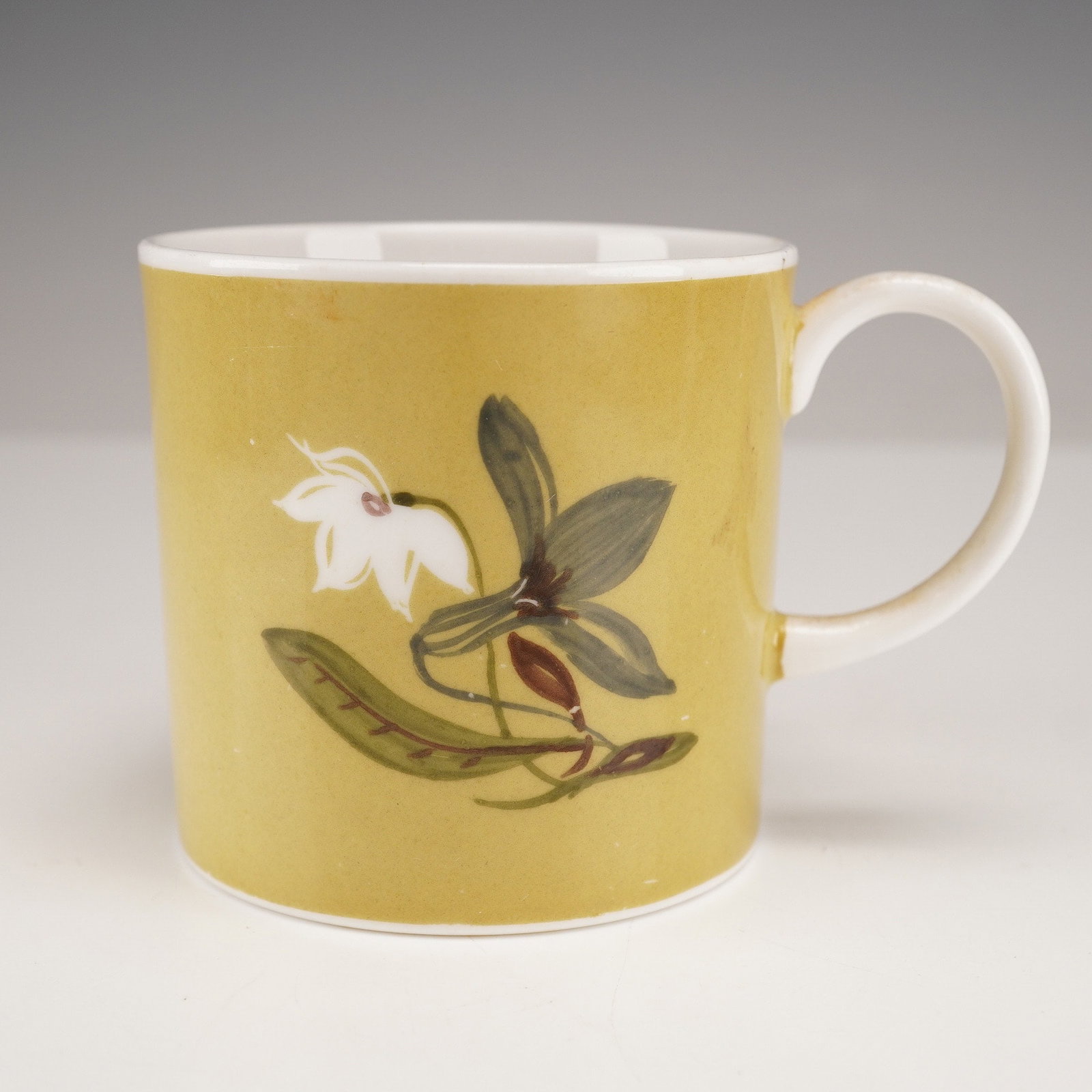 A Wedgwood Susie Cooper 'Flower Motif' Bone China Mug: A Wedgwood Susie Cooper 'Flower Motif' bone china mug. This cylindrical mug features a matte mustard yellow-ochre exterior glaze and a contrasting white interior, handle, and rim. The front is adorned