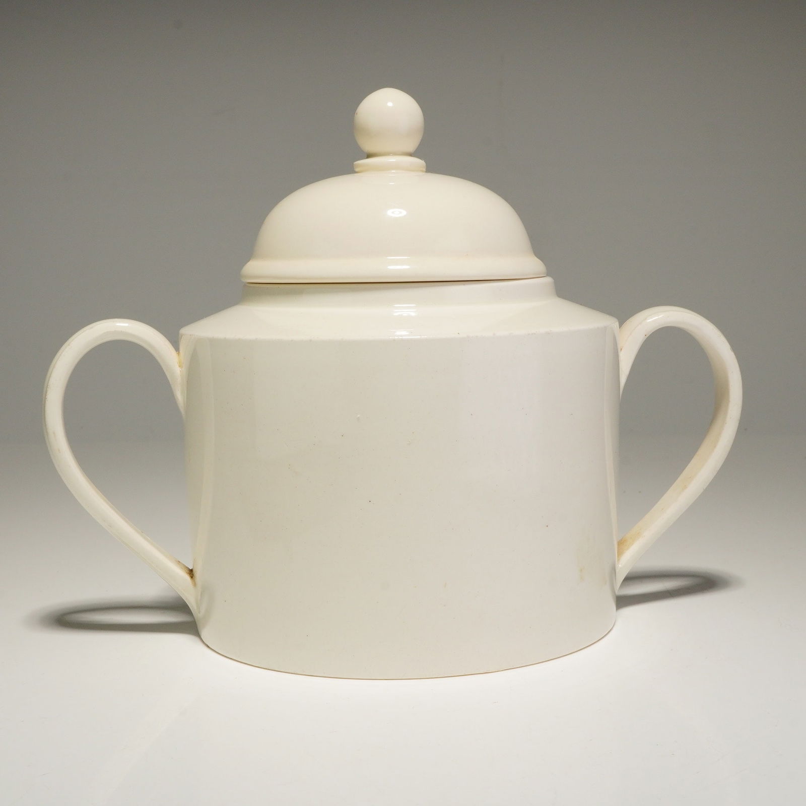 Wedgwood Double Handled Sugar Bowl with Lid: A Wedgwood bone china double handled sugar bowl with lid. This ceramic piece features a minimalist design with a cylindrical body and two loop handles. It is finished in an off-white glaze and include