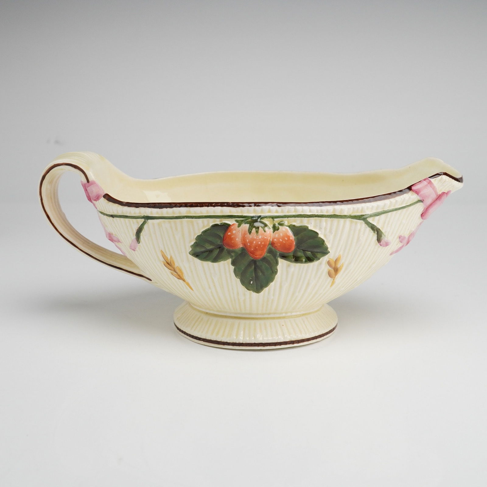 Wedgwood Gravy Boat with Fruit and Bow Motifs: This Wedgwood gravy boat is constructed from cream-colored earthenware featuring a reeded texture and molded, hand-painted decoration. The exterior is adorned on each side with a cluster of three stra