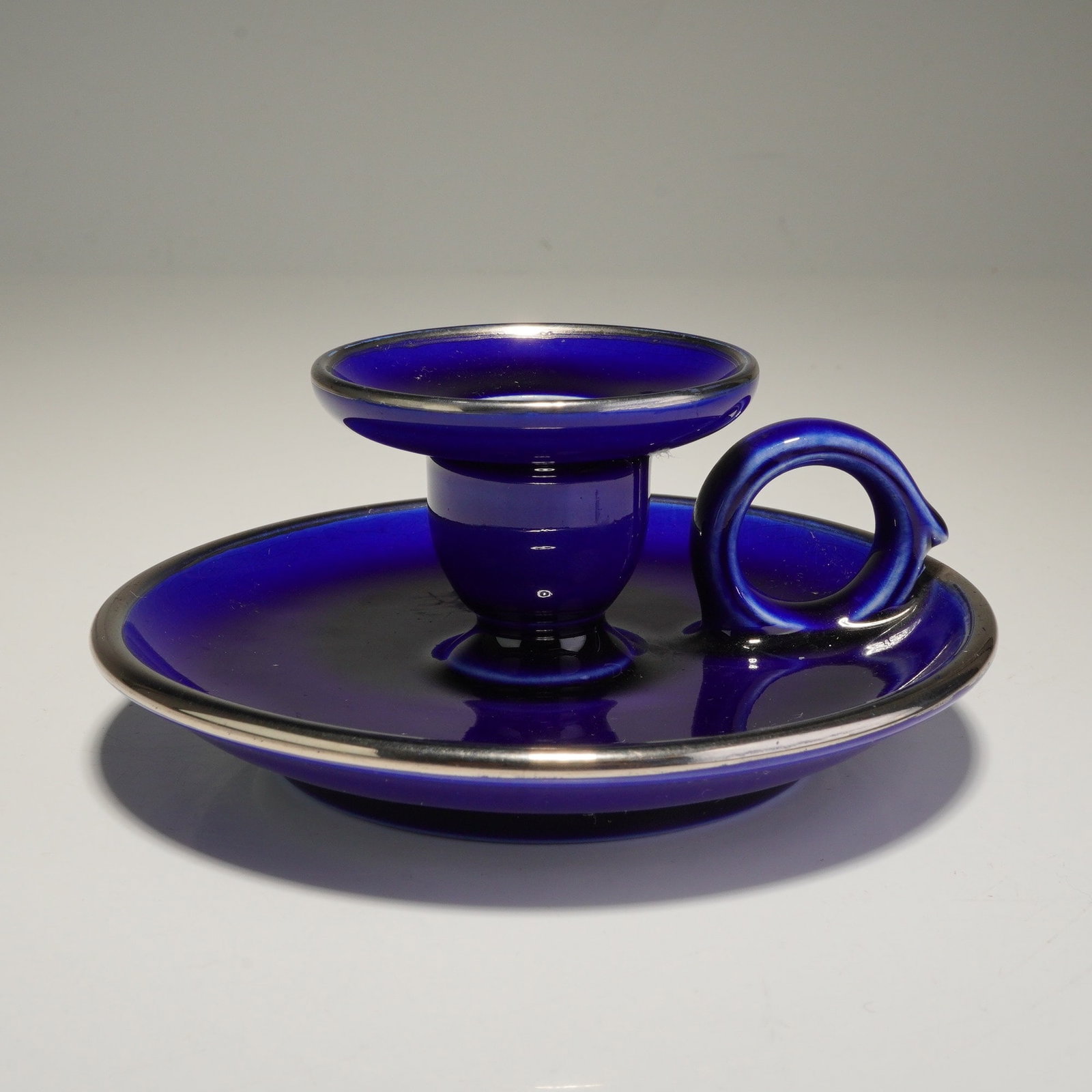 Wedgwood Cobalt Glazed Chamberstick: This ceramic chamberstick is finished in a deep cobalt blue glaze, consistent with a Wedgwood cobalt glazed chamberstick. The piece features a circular saucer base and a central candle cup, both accen