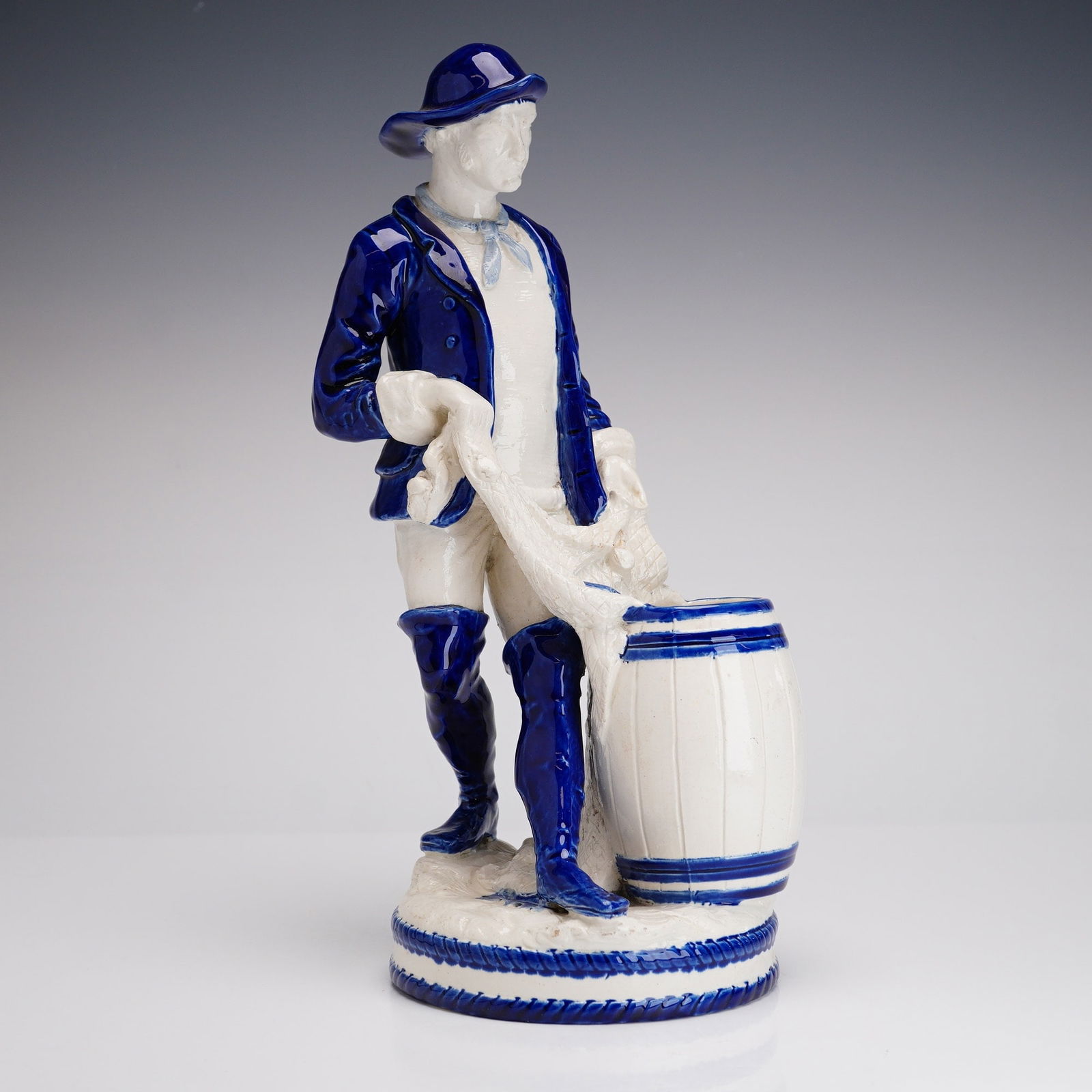 Royal Dux Bohemia Porcelain Fisherman Figurine with Wedgwood Blue Detailing: This molded porcelain figurine by Royal Dux Bohemia depicts a standing male fisherman or sailor in a traditional style. The piece is finished in a crisp white glaze with vibrant cobalt blue accents, c