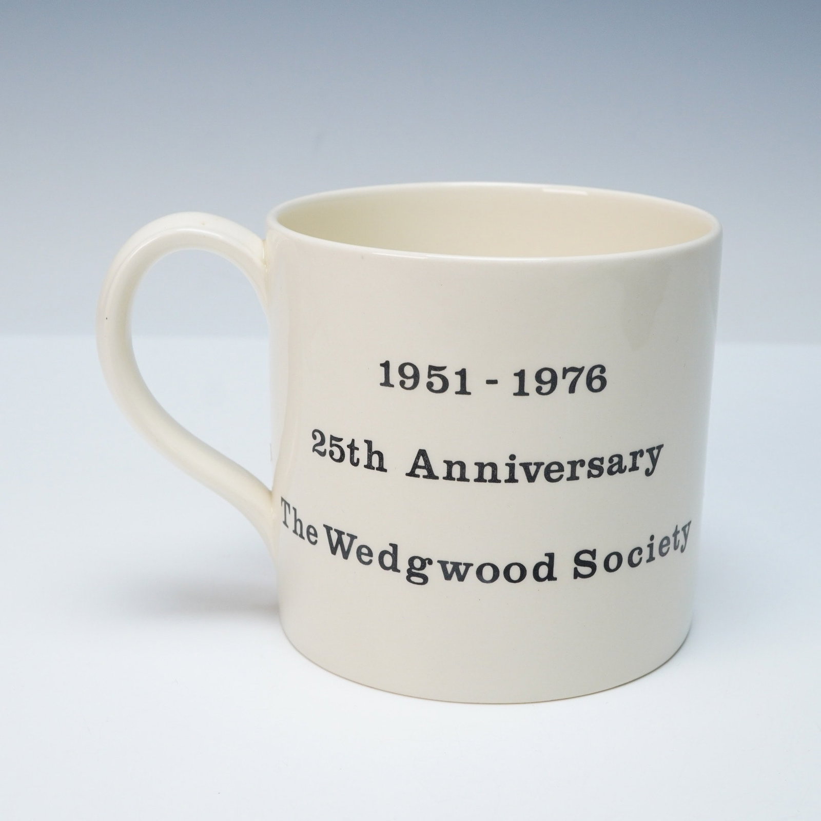 A Wedgwood Society 25th Anniversary Commemorative Mug: A cream-colored, glazed ceramic mug with a single handle by Wedgwood. The front is decorated with black printed text reading "1951 - 1976", "25th Anniversary", and "The Wedgwood Society". The reverse