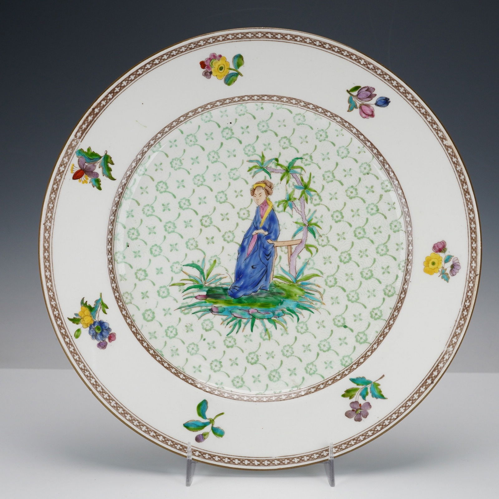 A Wedgwood Enameled Chinoiserie Plate: A Wedgwood enameled plate featuring decorations in a Chinoiserie style, depicting a seated figure in blue robe beneath a tree, surrounded by foliage against a light green diaper pattern ground in the