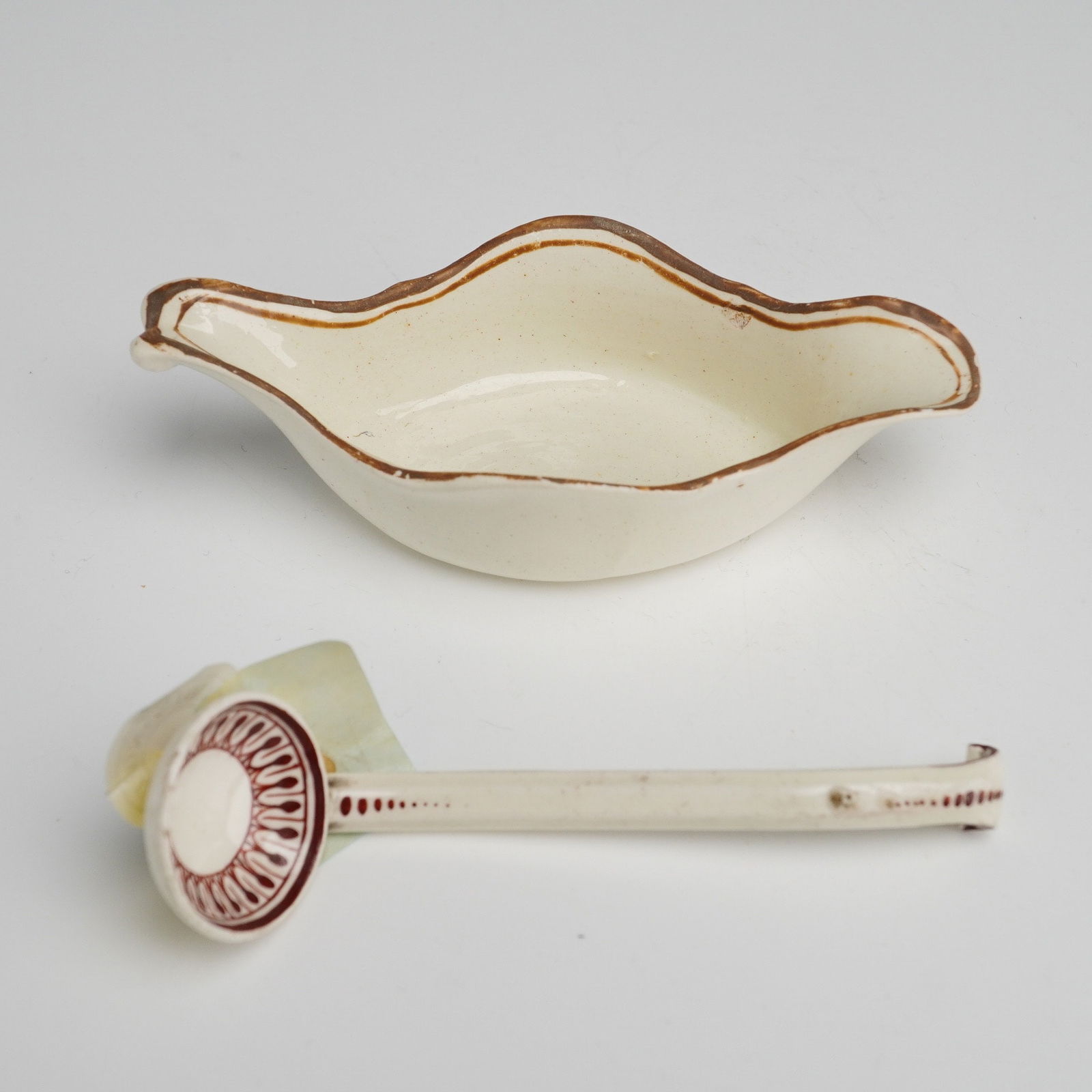 A Two-Piece Wedgwood Miniature Sauce Boat and Ladle: A two-piece Wedgwood miniature creamware set comprising a boat-shaped vessel and a matching long-handled ladle. The items are finished with a cream-colored glaze. The vessel features a simple brown-ba