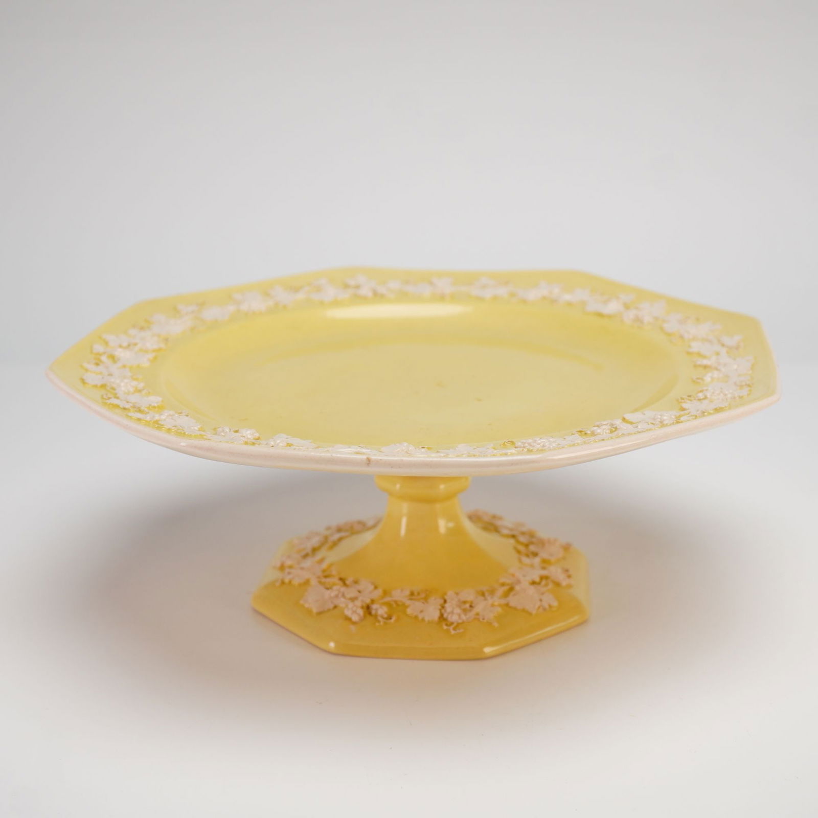 Wedgwood Etruria Embossed Queen's Ware Yellow Octagonal Pedestal Cake Stand: A Wedgwood Embossed Queen's Ware octagonal pedestal cake stand featuring a pale yellow glazed ground. The piece is decorated with a grapevine motif in raised relief, consisting of applied white bas-re
