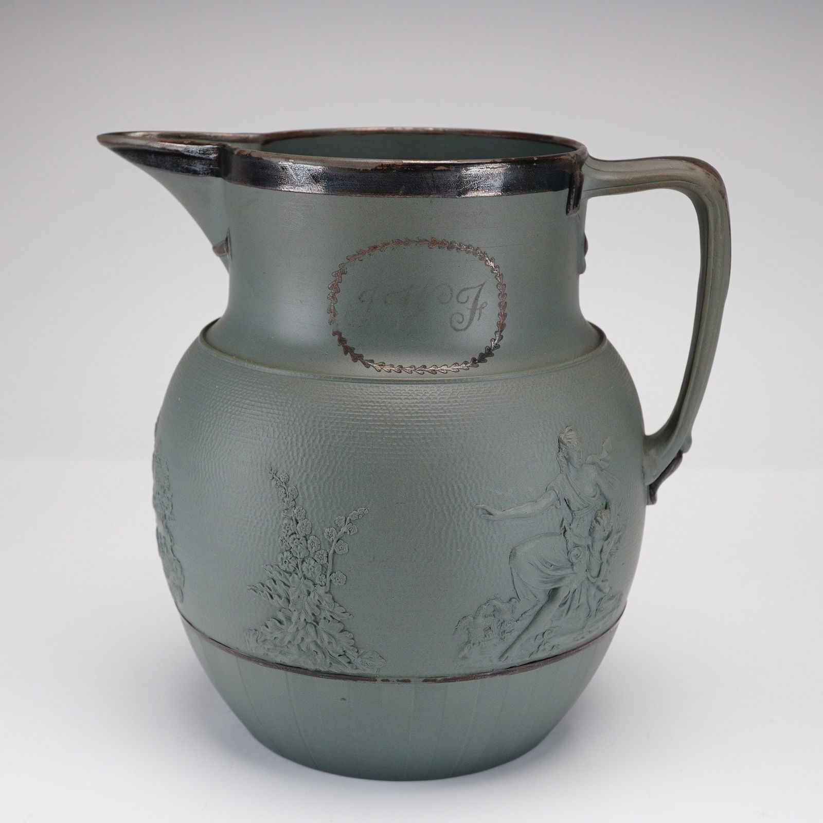A Wedgwood Drabware Relief Molded Stoneware Jug: A Wedgwood drabware relief molded stoneware jug, England, circa 1820. The vessel features a sage green stoneware body decorated with applied relief scenes depicting classical figures, trees, and flora