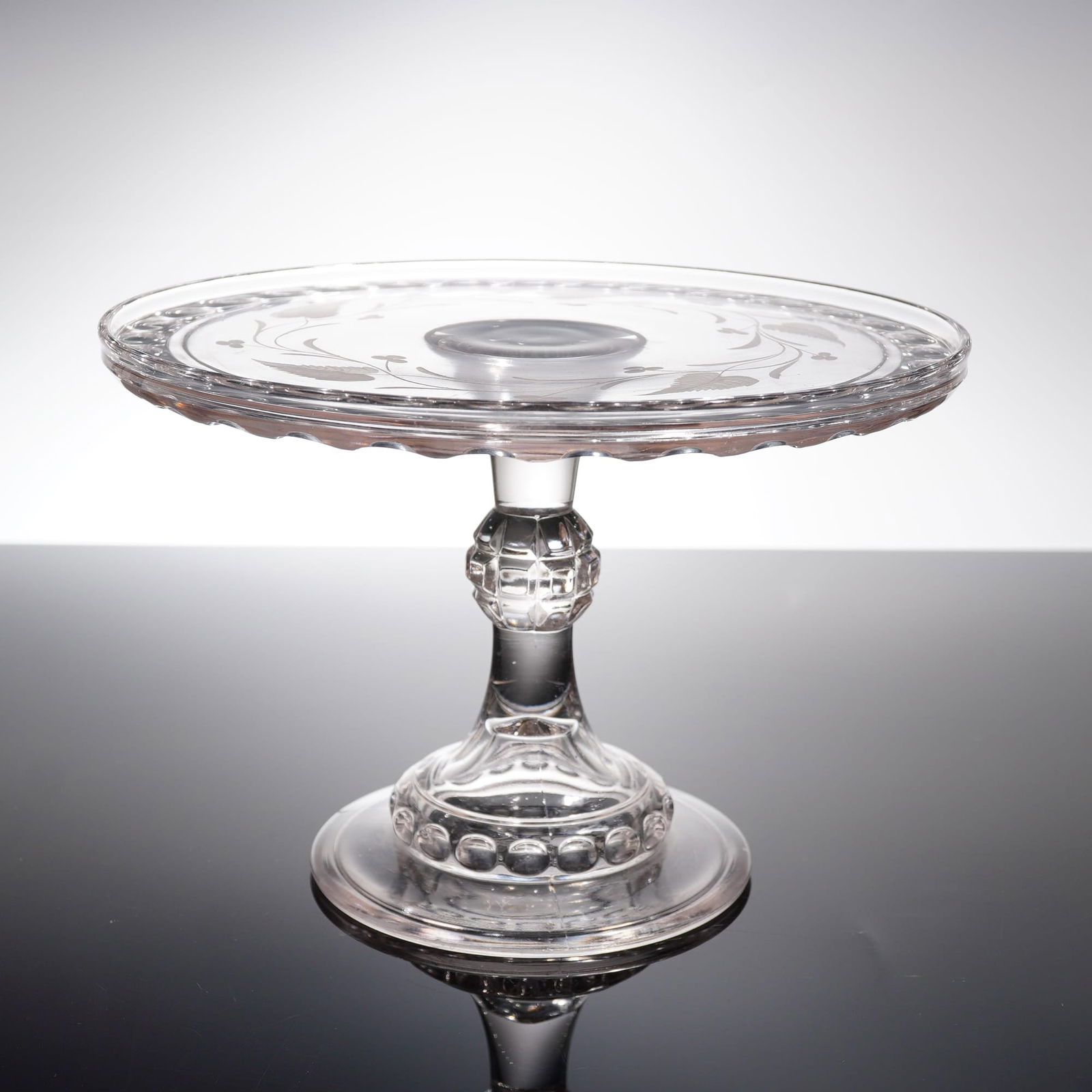Early American Pressed Glass Cake Stand: A single pedestal dish, commonly known as an Early American Pressed Glass cake stand. The object is constructed of clear glass, featuring a circular plate supported by a stem and circular foot. The te