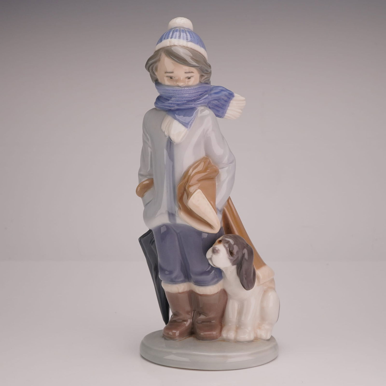 Lladró Porcelain Figurine, Winter Boy with Dog #5220: A Lladró porcelain figurine titled Winter (model 01005220), depicting a young boy prepared for cold weather. The figure is dressed in a light gray overcoat, blue trousers, and brown boots, accented b