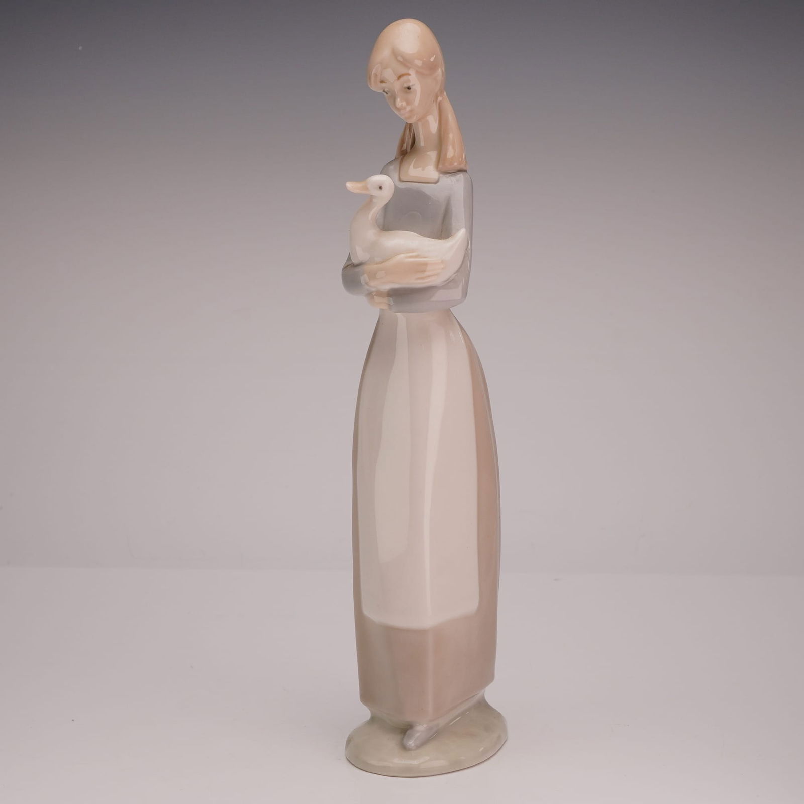 Nao by Lladró Porcelain Figurine, Shepherd Girl with Goose: This slender, stylized Nao by Lladró porcelain figurine depicts a young shepherd girl standing and cradling a large white goose in her arms. The figure is modeled with long, soft brown hair and wears