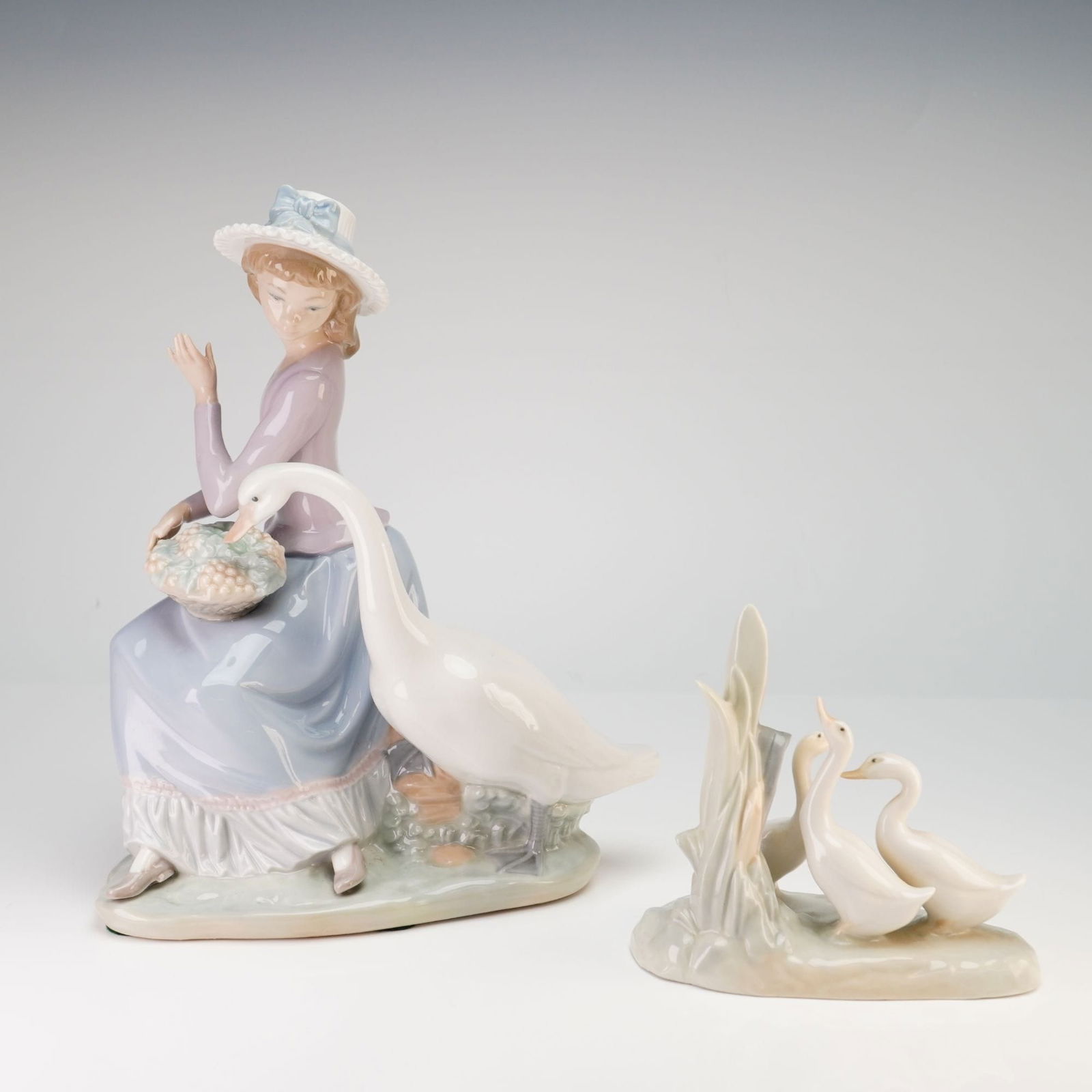 2pc Lladro and Nao by Lladro Porcelain Avian Figurines: This lot consists of two glazed porcelain figurines manufactured in Spain. The first figurine is a Lladro piece identified as Goose Trying to Eat, model number 5034, designed by Vicente Martinez. The