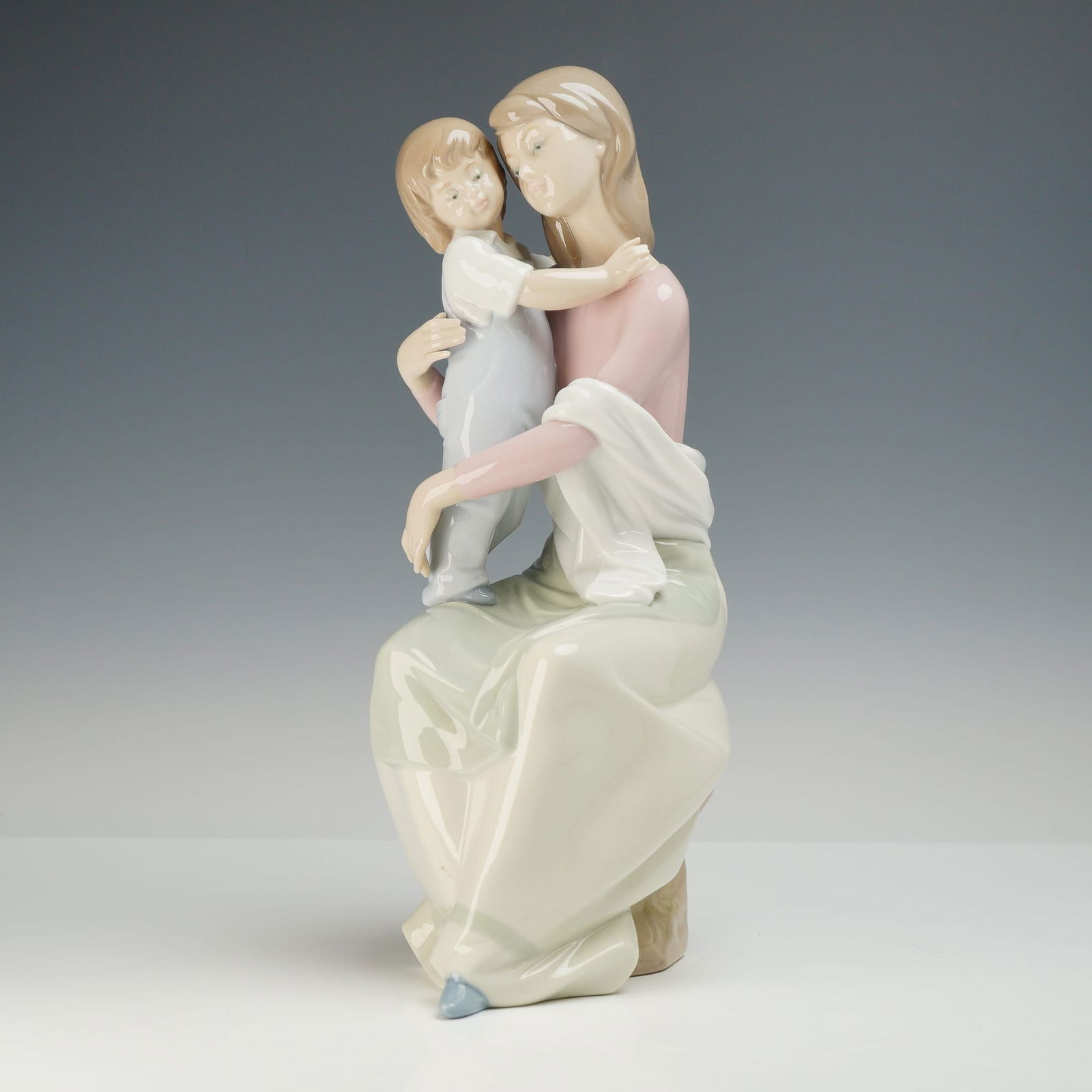 Lladró A Mother's Love Glazed Porcelain Figurine 6634: This glazed porcelain figurine, titled A Mother's Love, is produced by Lladró. The piece depicts a seated woman in a pink bodice and pale green skirt holding a young child wearing light blue overalls