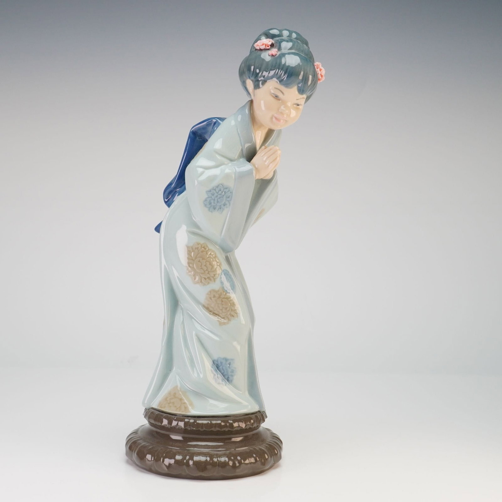 Lladro Sayonara Porcelain Figurine Model 4969: A Japanese woman in a traditional bowing posture with hands held together. She wears a light blue kimono featuring a floral pattern in tan and blue, complemented by a dark blue obi. Her dark hair is a