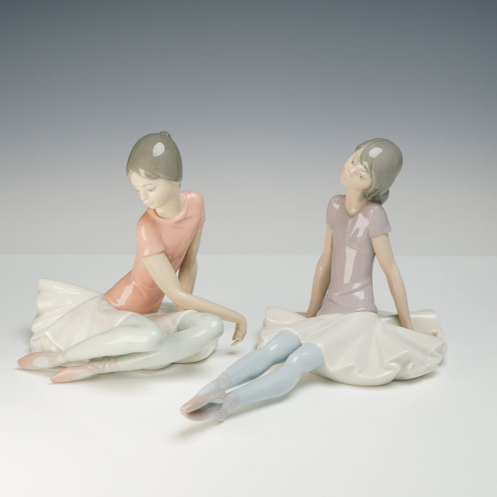 2pc Lladro Porcelain Ballerina Figurines, Shelley & Phyllis: This pair of glazed porcelain figurines is manufactured by Lladro in Spain. The set features two young ballerinas in seated, restful poses. One figure, identified as Shelley (model 1357), is depicted