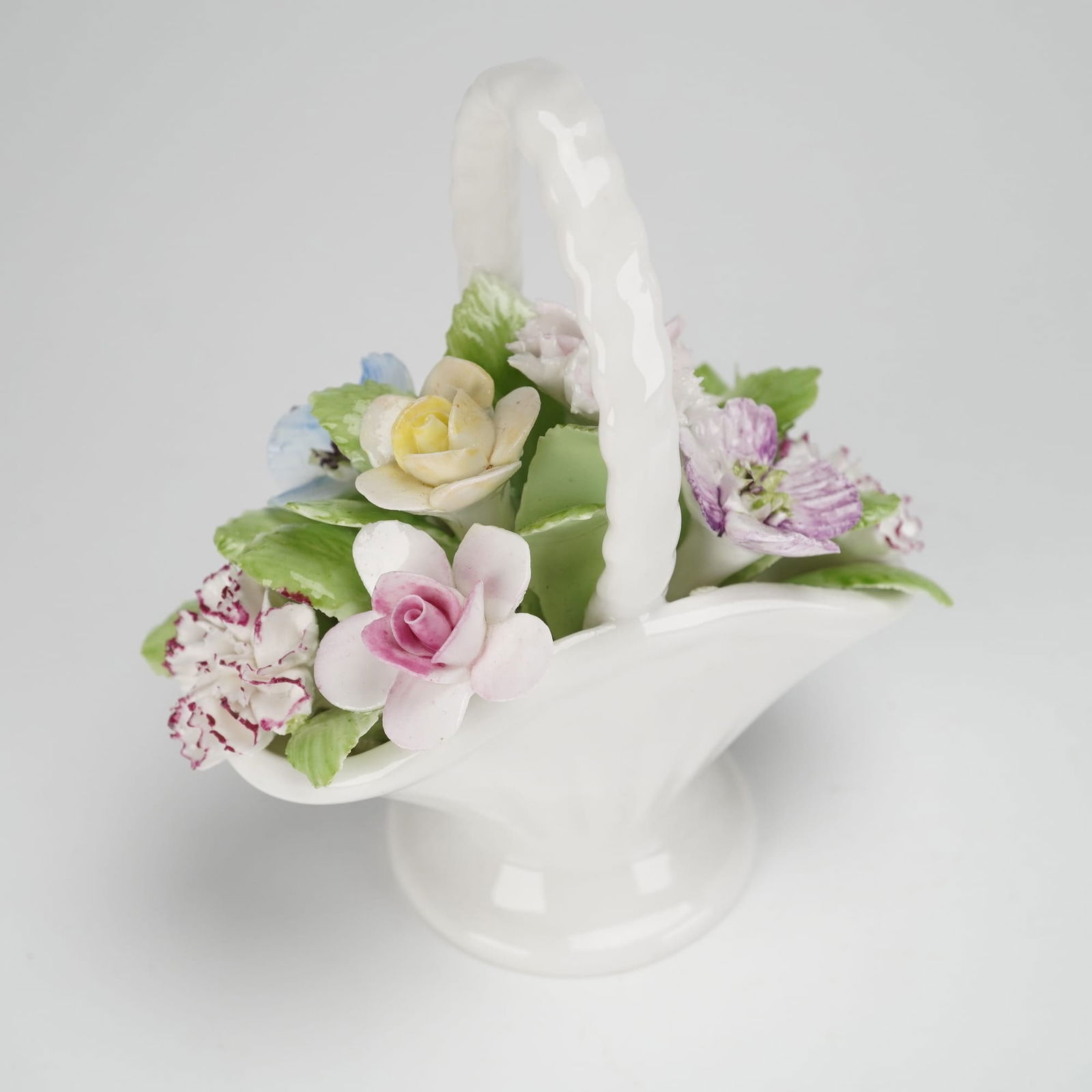 Royal Doulton Bone China Figurine, Handled Basket of Flowers: The figurine features a stylized, fan-shaped white basket with a braided arched handle, resting on a circular foot. The basket contains an arrangement of hand-modeled and applied ceramic flowers, incl