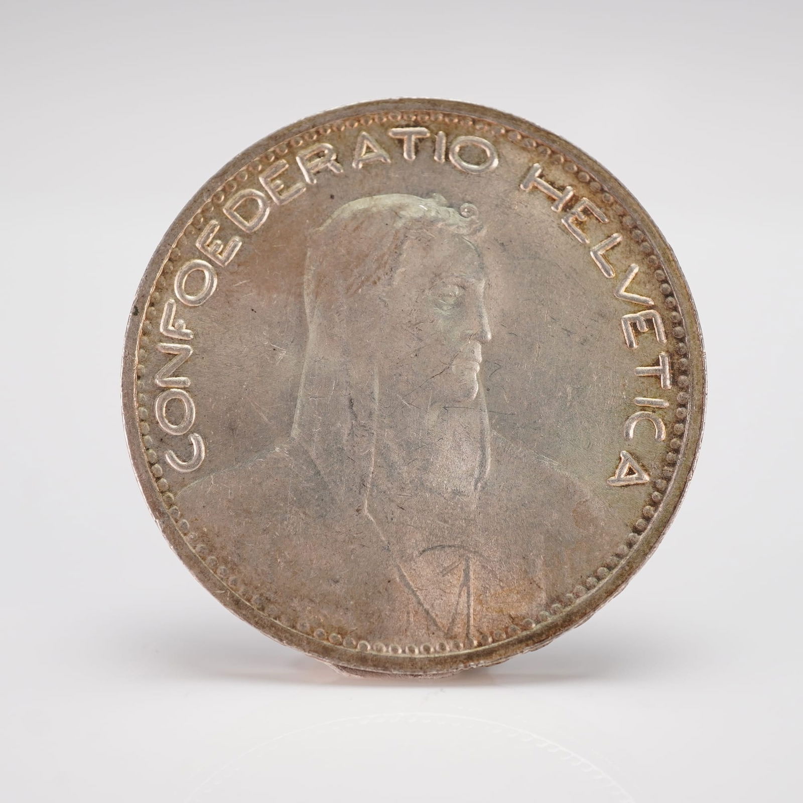 1923 B Switzerland 5 Francs Silver William Tell Coin: A 1923 B Switzerland 5 Francs Silver William Tell coin. This circular coin is crafted from a silvery-toned metal, exhibiting significant darkening and tarnishing consistent with age and environmental