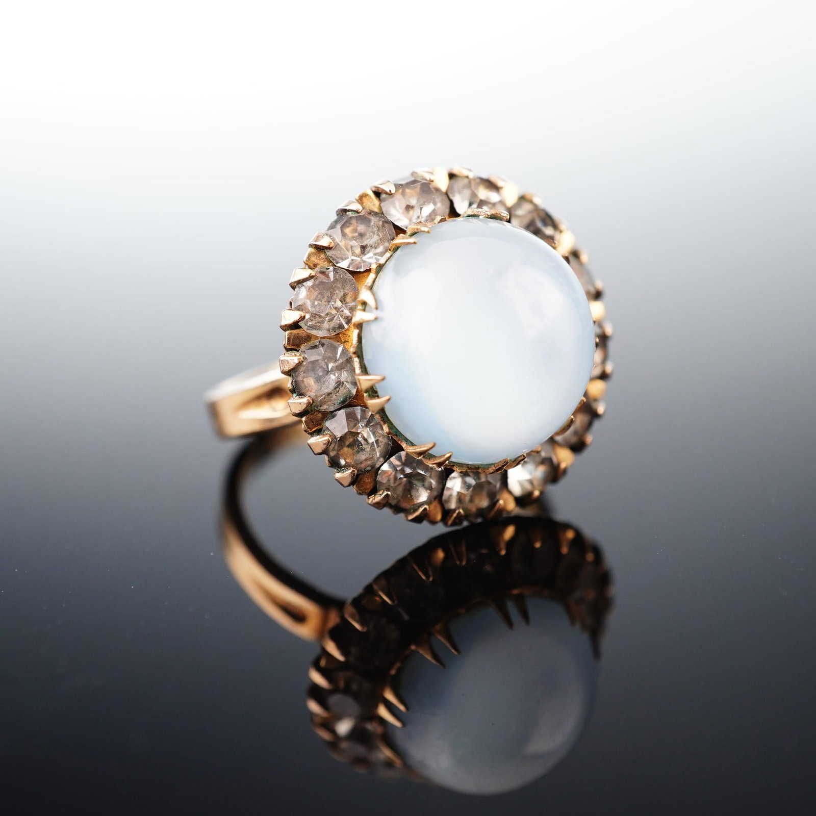 Clarks & Coombs 14K Rolled Gold Plate Moonstone Cluster Ring: A cocktail ring by Clarks & Coombs featuring a large, round cabochon moonstone center stone displaying a changeable white to pale blue luster. The center stone is encircled by a halo of round-cut clea