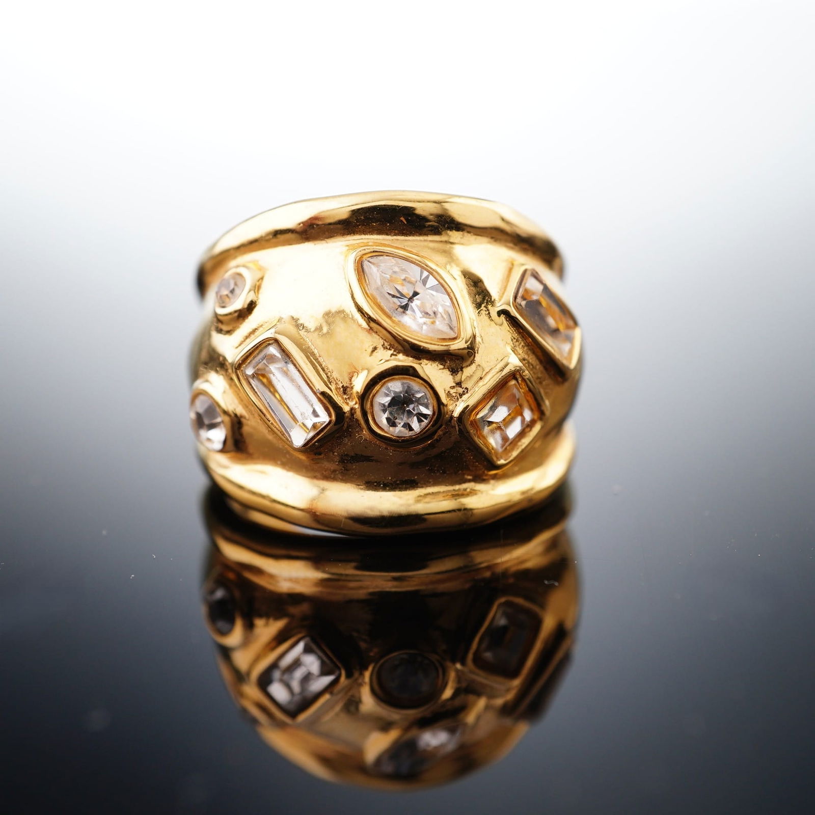 Gold Plated Clear Stone Inlaid Wide Band Ring: This gold plated stone inlaid ring features a bold, sculptural aesthetic featuring a polished, organic melted texture. The wide, convex band is inset with multiple colorless multi-cut stones in variou