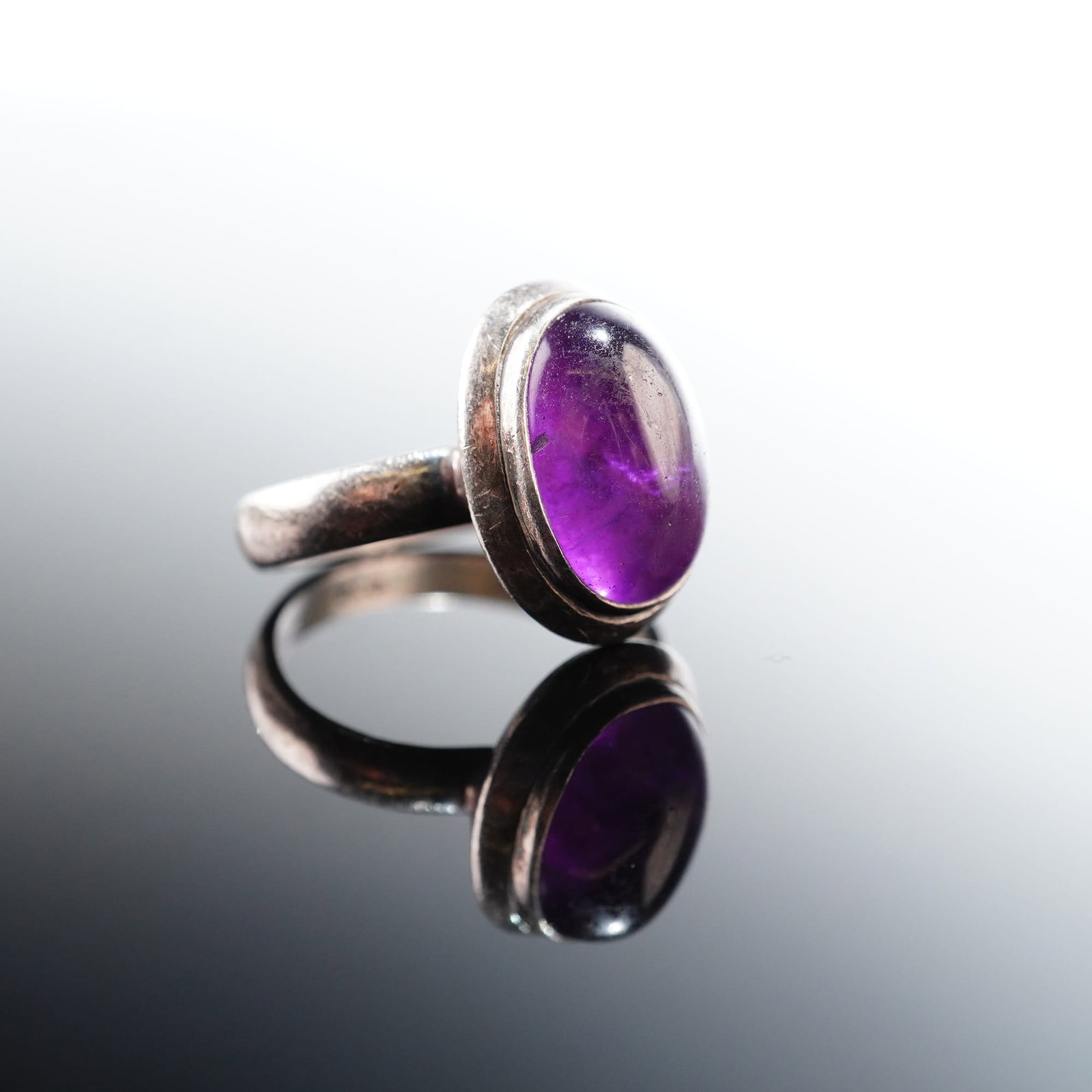 Oval Amethyst Cabochon Sterling Silver Ring: Sterling silver ring set with an oval-shaped amethyst cabochon. The polished silver mounting frames the smooth, domed gemstone in a classic setting, highlighting the rich purple hue of the stone.