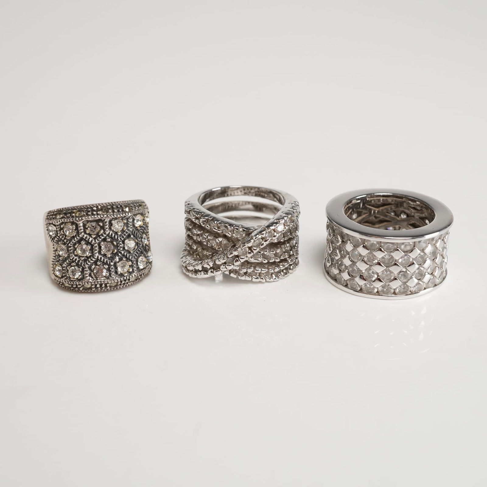 A Trio of Silver Fashion Rings: Comprises coordinated silver-tone band rings featuring varied dimensional profiles and decorative surface treatments, including pavÃ©-set clear rhinestones, geometric lattice-style settings, and