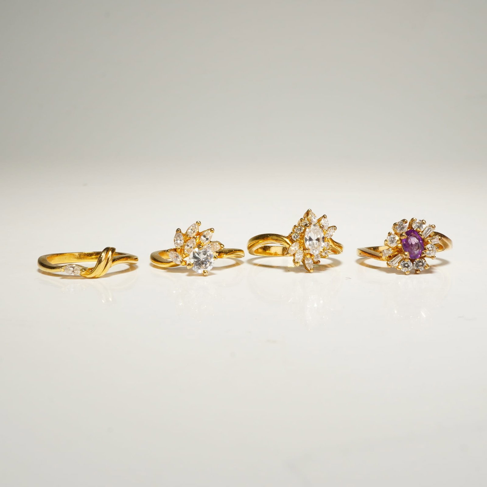 4pc Gold-Tone Rhinestone Fashion Rings: This lot consists of four yellow gold-tone rings in various designs. The first ring is a bypass style featuring a marquise-cut colorless stone and a twisted band. The second is an asymmetrical