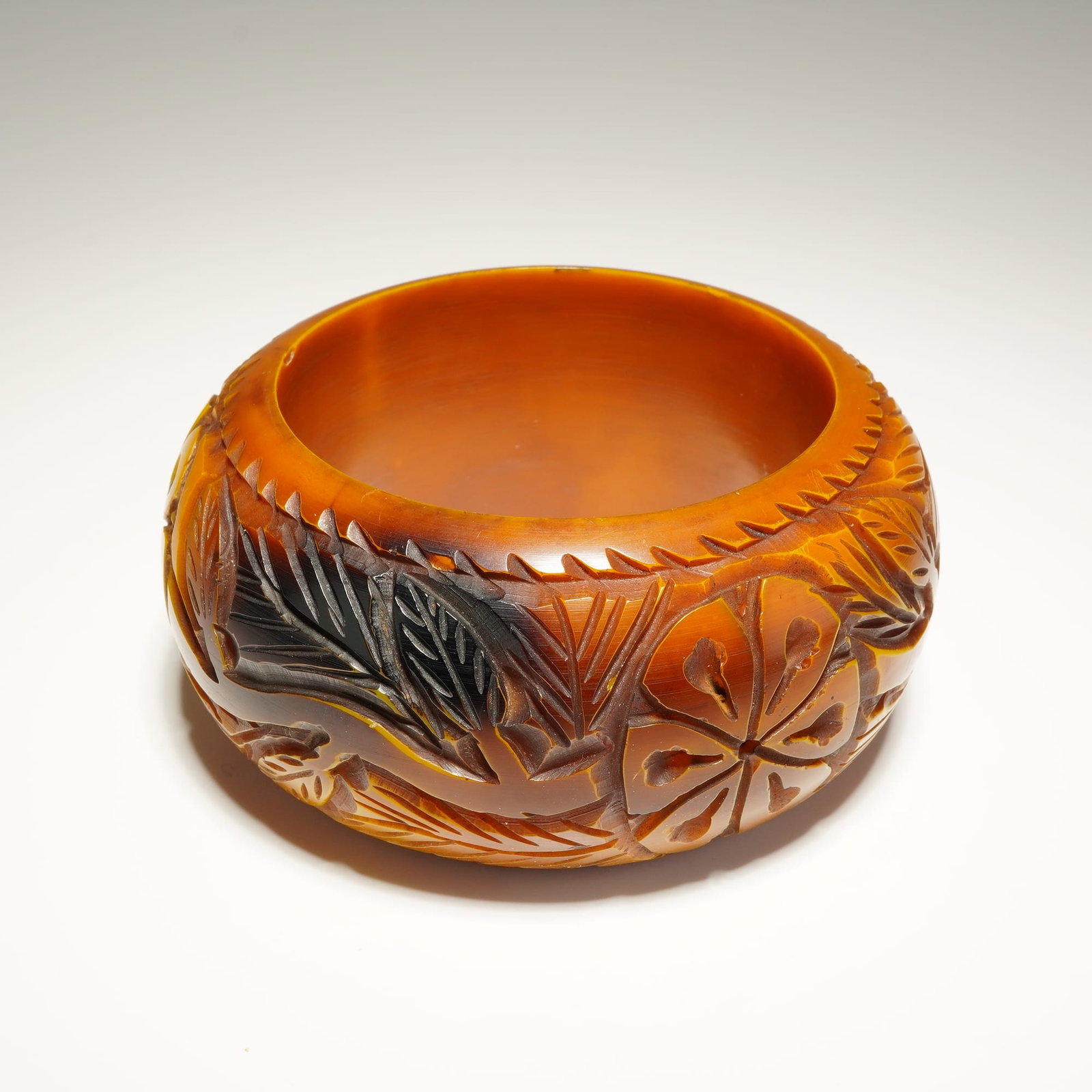 Carved Butterscotch Bakelite Floral Motif Bangle Bracelet: This wide, vintage bangle featuring a continuous, deeply carved relief motif of stylized flowers and foliate patterns encircling the exterior. The material exhibits a rich color transition from bright