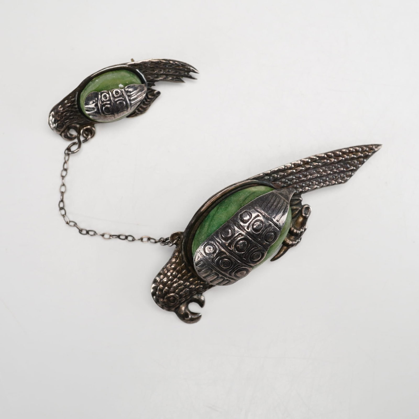 Mexican Sterling Silver and Green Stone Double Parrot Brooch: This sterling silver bird brooch features two stylized parrots connected by a silver safety chain. Each bird is designed with an oval green stone body set within a textured silver frame that forms the