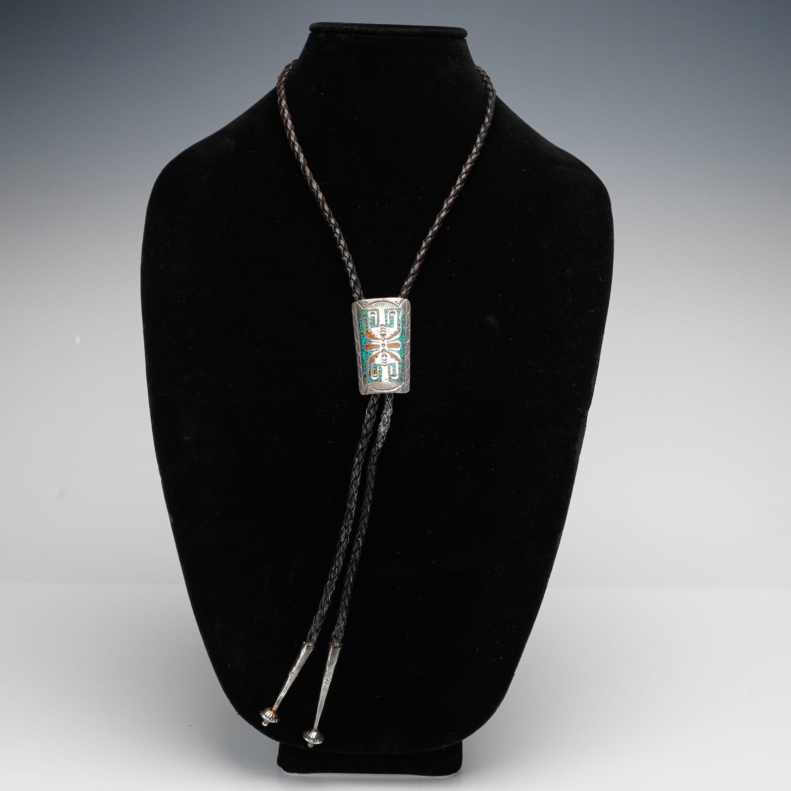 Bennett Sterling Silver Turquoise and Coral Inlay Bolo Tie: A bolo tie featuring a rectangular sterling silver slide with rounded corners. The slide is embellished with a symmetrical, abstracted central motif in silver, surrounded by turquoise and coral chip i