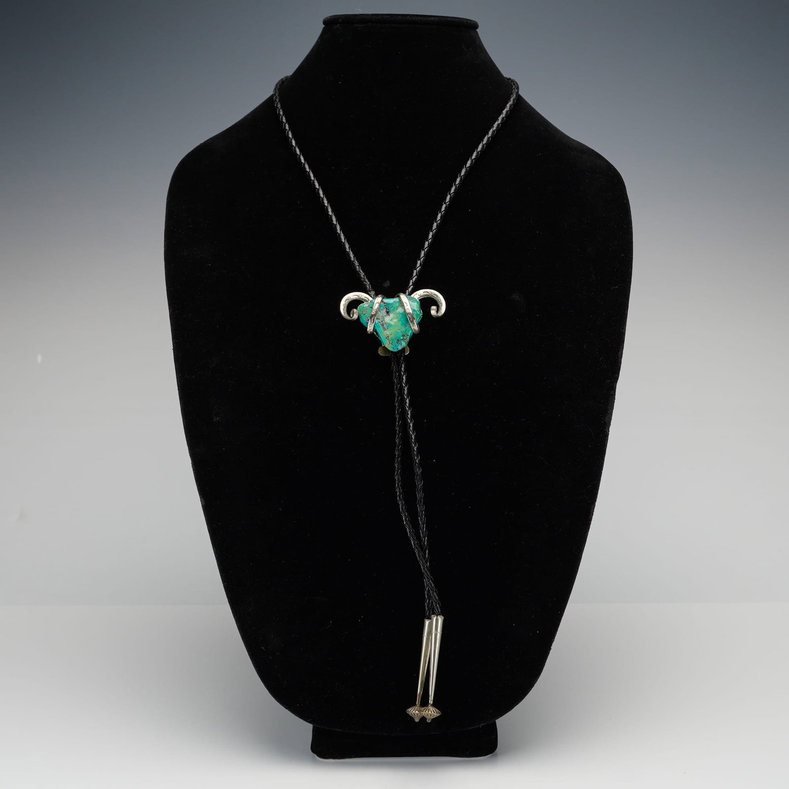 Turquoise and Silver Metal Ram Bolo Tie: The bolo tie features a large matrixed turquoise stone embraced by silver tone metal elements that wrap around the stone and finish in curved designs reminiscent of ram horns, decorated with stamped a