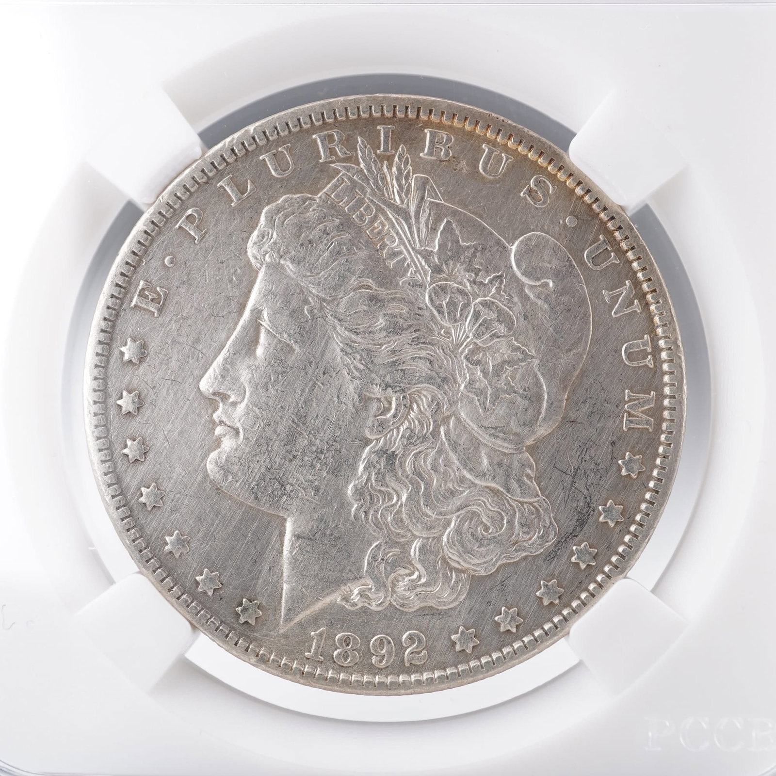 1892-O United States Morgan Silver Dollar: The obverse of this 1892 O Morgan Silver Dollar features the profile of Liberty wearing a Phrygian cap adorned with cotton and wheat, symbolizing freedom and agricultural strength. Surrounding her ima