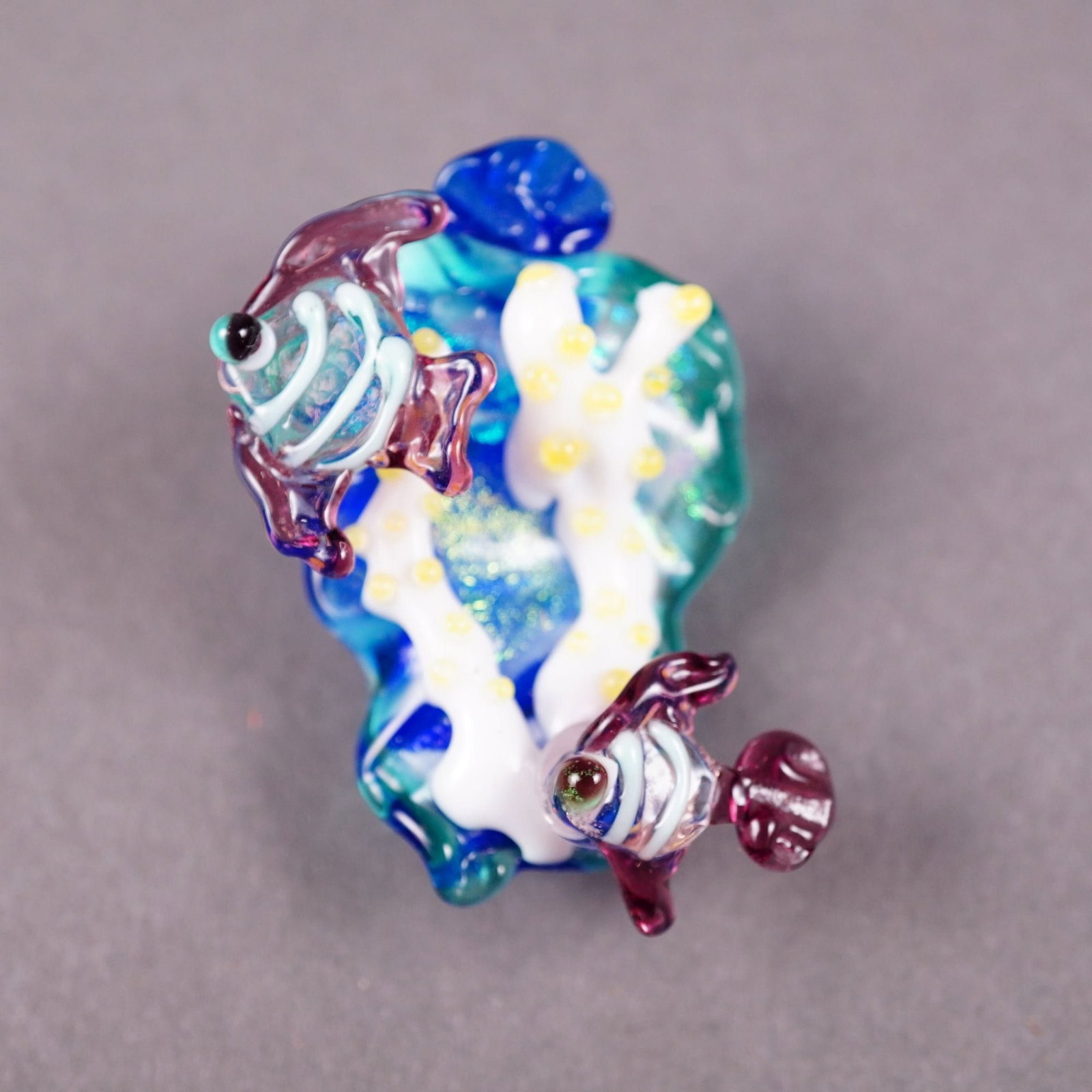Studio Artist Lampwork Glass Aquatic Fish Button: A hand-crafted studio glass button featuring an intricate lampwork aquatic scene. The surface is expertly sculpted with two amethyst-toned striped fish swimming amidst a white branching coral reef acc