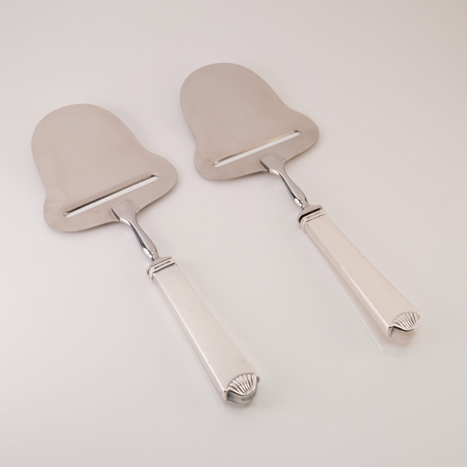 A Pair of Hans Hansen Haakon Pattern Sterling Silver and Stainless Steel Cheese Planes: A pair of Danish modern cheese planes by Hans Hansen in the Haakon pattern. Each piece features a sterling silver handle with a classic shell-motif terminal and a stainless steel slicing blade. The ha
