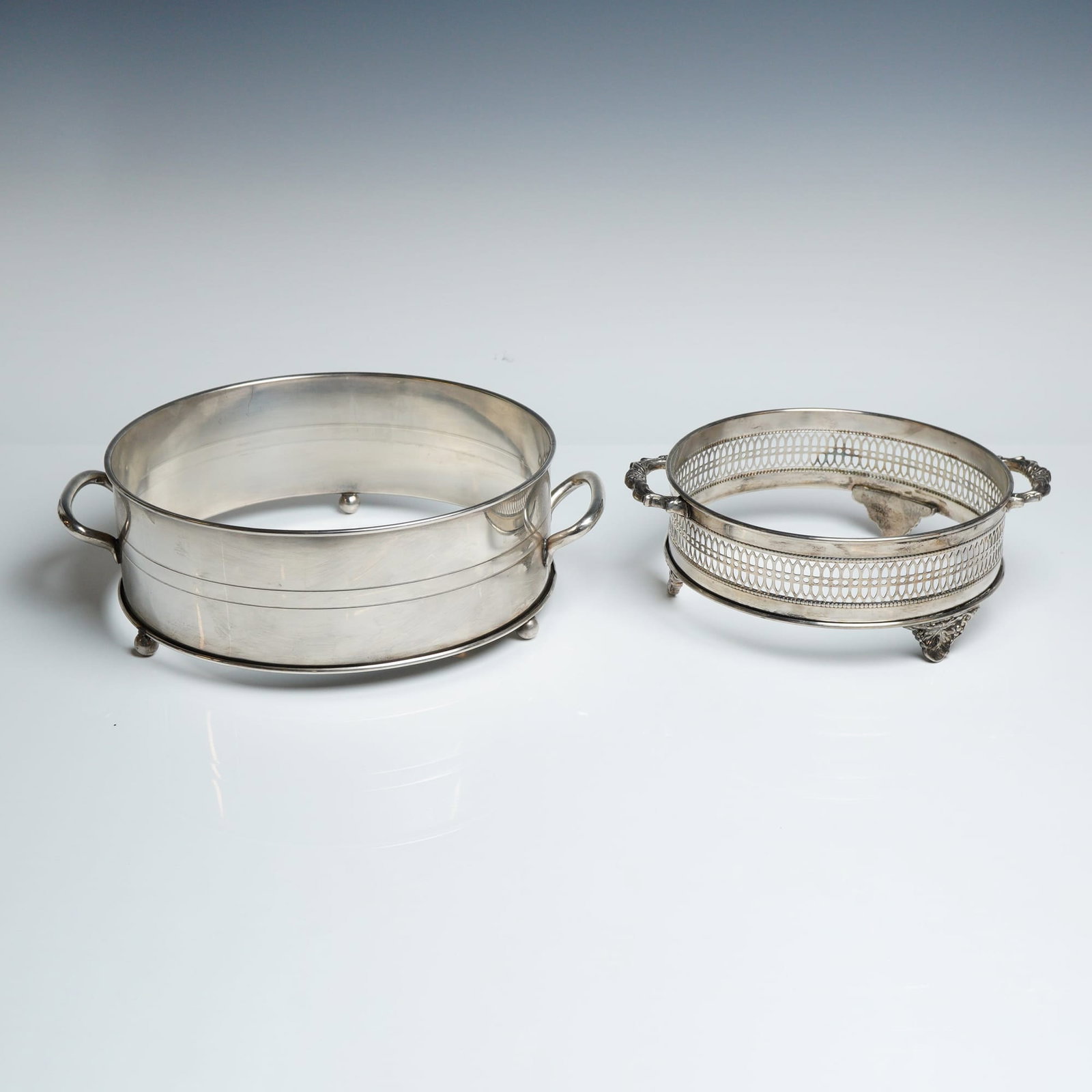 Two Silverplated Casserole Dish Holders: Two silverplated casserole dish holders of varying sizes and styles. The larger holder is of a plain, circular design with two parallel incised lines around the body, featuring two simple D-shaped han