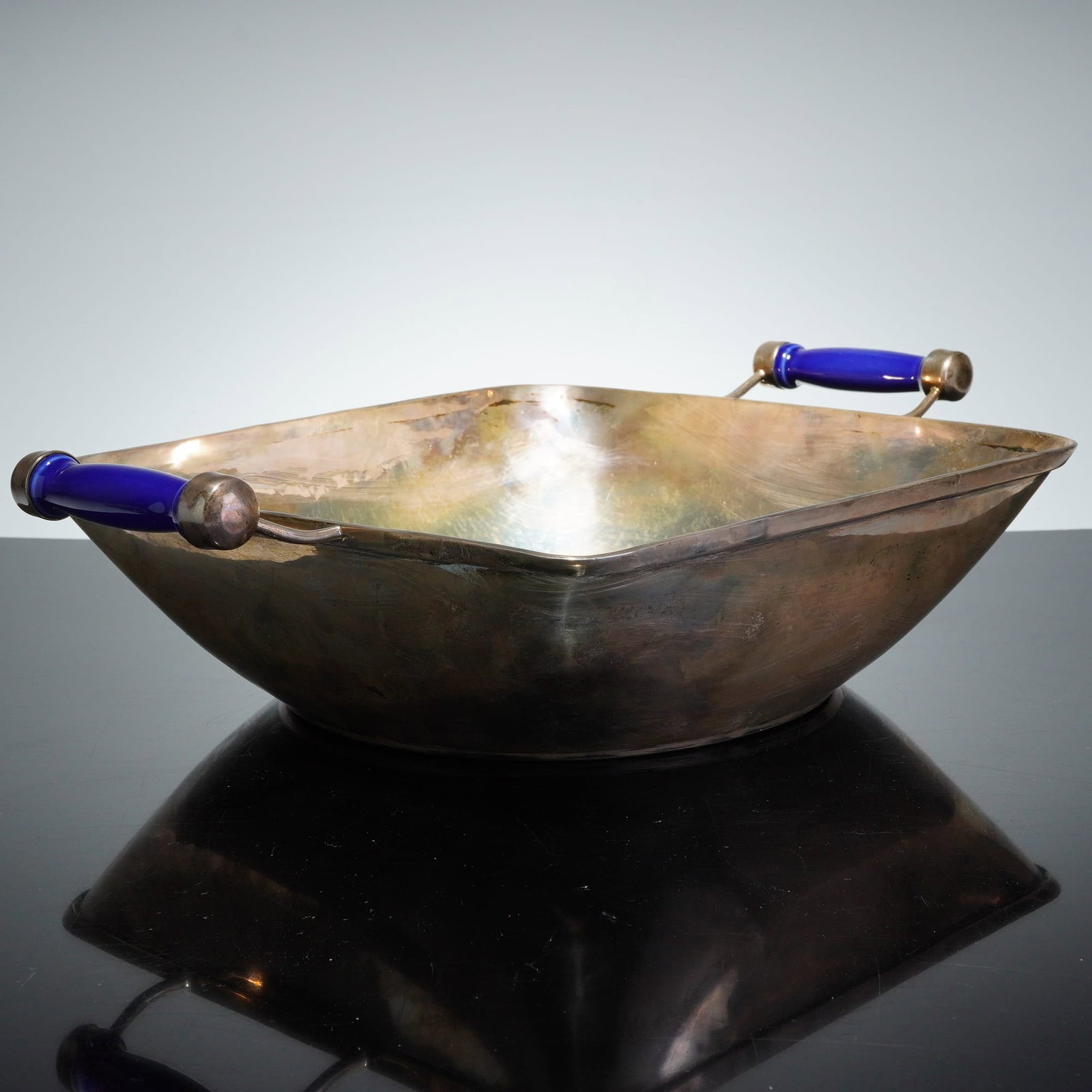 Mid-Century Modern Sterling Silver Serving Bowl with Cobalt Handles: A mid-century modern sterling silver serving bowl featuring a rounded square form with flared sides and a subtle hammered interior finish. The dish is accented by two cobalt blue glass handles set in