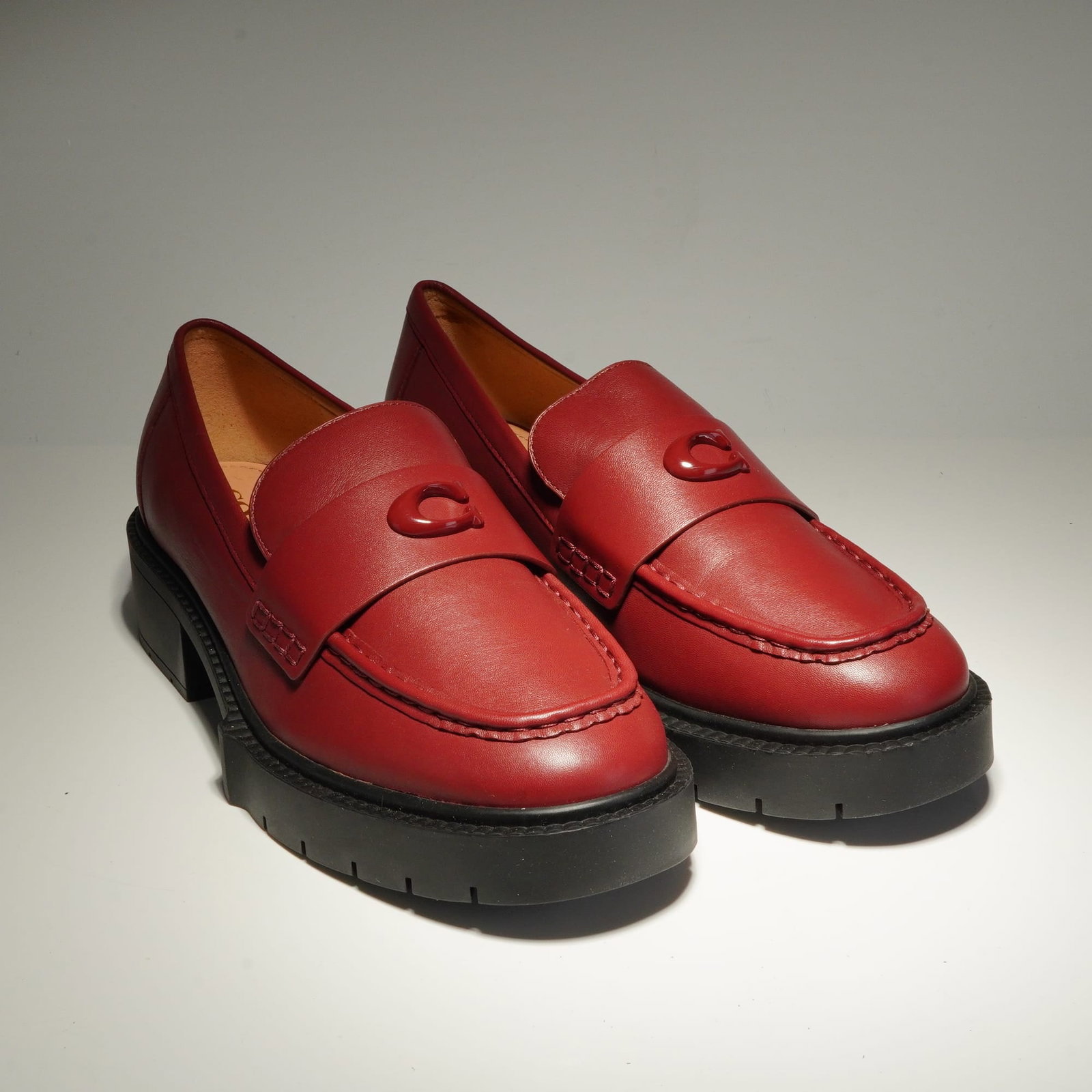 Coach Women's Leah Loafers in Wine Leather: A pair of Coach Leah loafers featuring ruby red leather uppers. The design incorporates a moc-toe construction with a tonal Coach C emblem situated on the vamp strap. The footwear is built upon substa