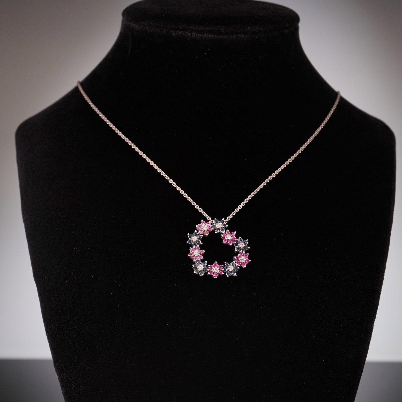 18K White Gold Diamond and Sapphire Floral Heart Pendant Necklace: This white gold heart-shaped pendant is outlined by flowers made from alternating blue and pink sapphires, each with a diamond center. The pendant hangs from a thin chain necklace. A tag near the clas