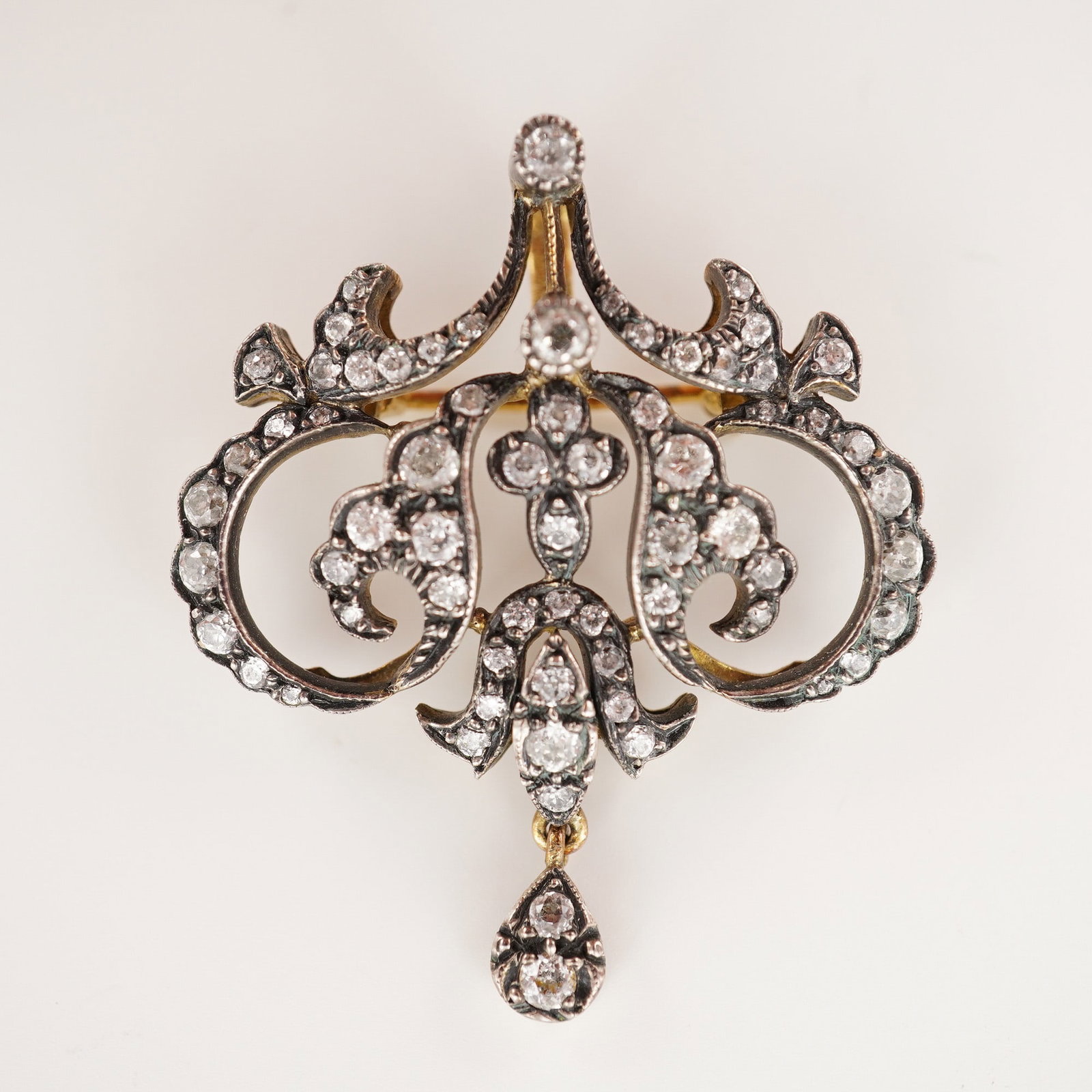 Diamond Foliate Scroll Brooch Pendant: Openwork scrolling foliate design set with numerous round-cut diamonds in graduated sizes, centered by a trefoil motif and suspending a pear-shaped diamond-set drop. Crafted in gold-toned metal and fi