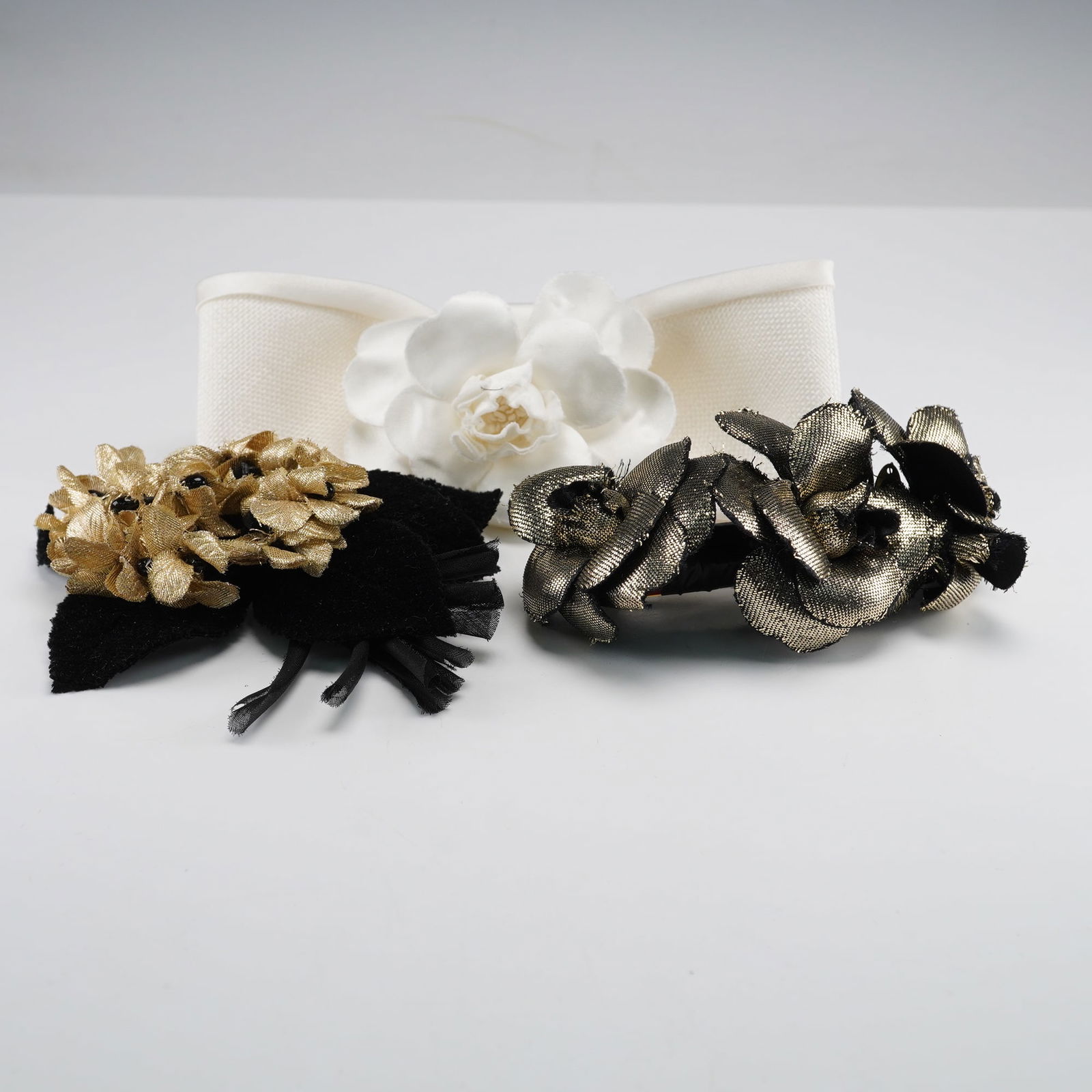 Three Chanel France Hair Barrettes: A group of three Chanel luxury hair accessories. Comprising a cream-colored textured fabric bow barrette adorned with a central white fabric camellia-like flower, featuring a gold-tone metal tag embos