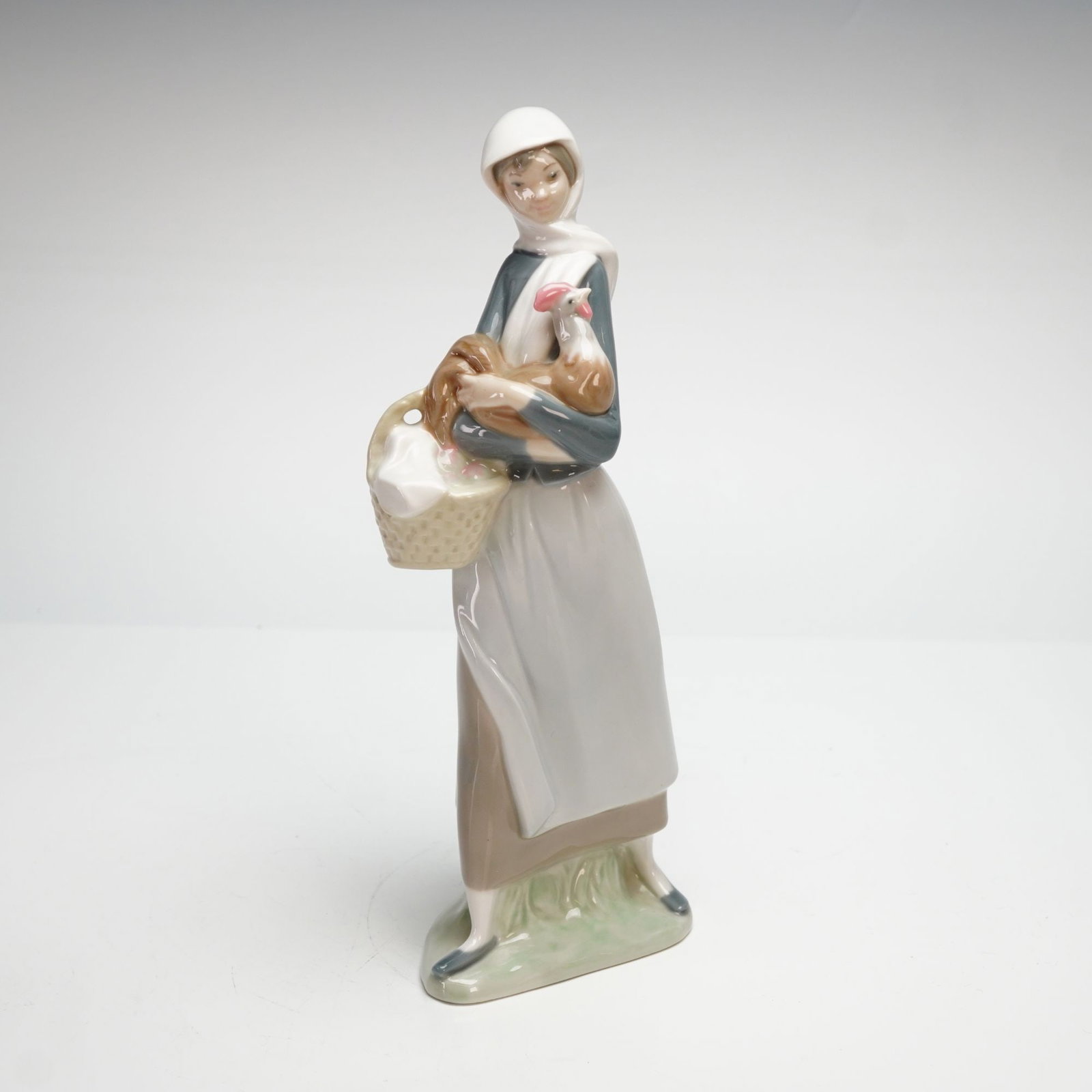 Lladro Porcelain Figurine, Girl with Cockerel 1004591: This polychrome glazed porcelain piece depicts a young woman in traditional rustic attire, wearing a white headscarf, a blue long-sleeved bodice, a long tan skirt, and a white apron. She is shown stan