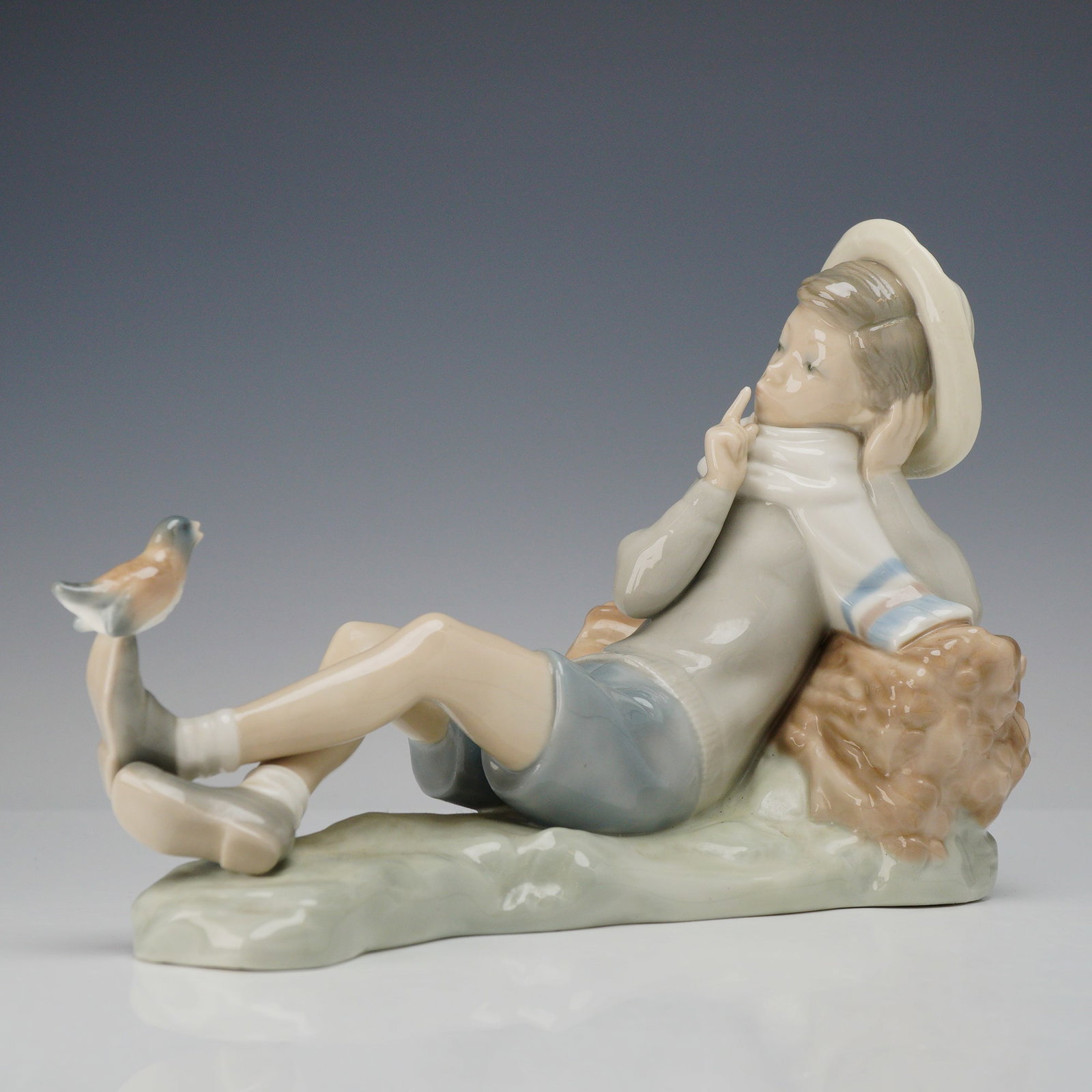 Lladro Porcelain Figurine, Shepherd with Bird 1004730: This Lladro glazed porcelain figurine is depicts a young boy reclining against a bundled bale of hay. He is dressed in a wide-brimmed hat, a white scarf with pale blue stripes, a gray-brown shirt, and