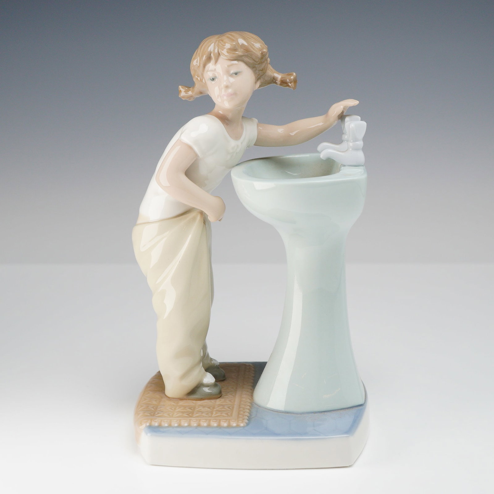 Lladro Figurine, Clean Up Time 1004838: A vintage Lladro glossy porcelain figurine. The charming scene depicts a young girl with pigtails, dressed in a white t-shirt and tan trousers, leaning over a light blue pedestal washbasin. She is pos