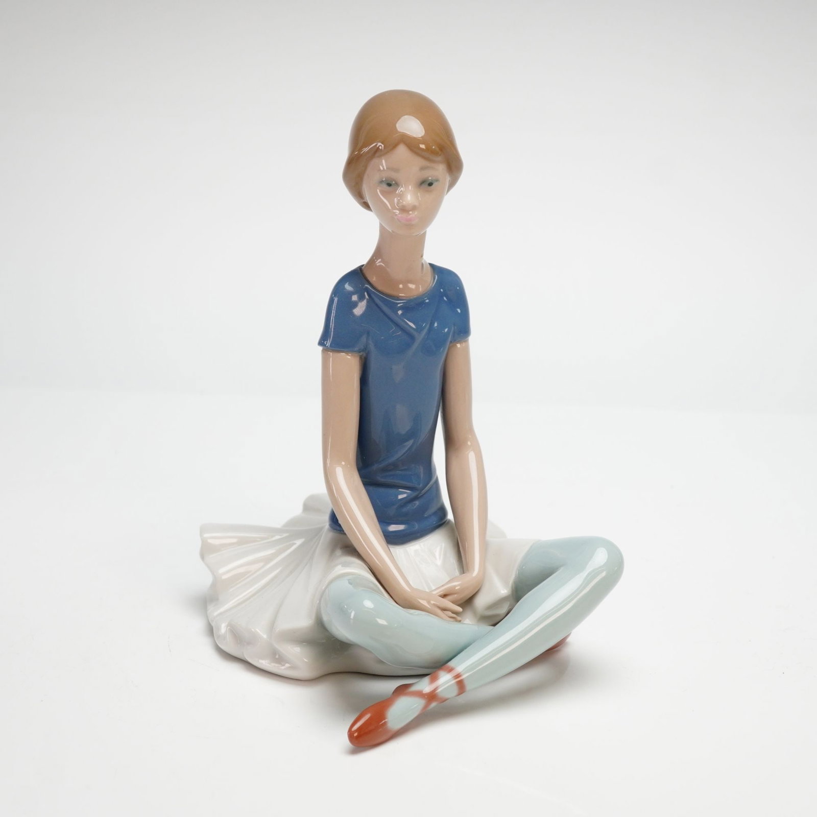 Lladro Porcelain Figurine, Beth 1001358: This glazed porcelain figurine depicts a young ballerina in a seated, cross-legged pose, wearing a short-sleeved blue top, a voluminous white tutu skirt, light blue tights, and reddish-brown ballet sl
