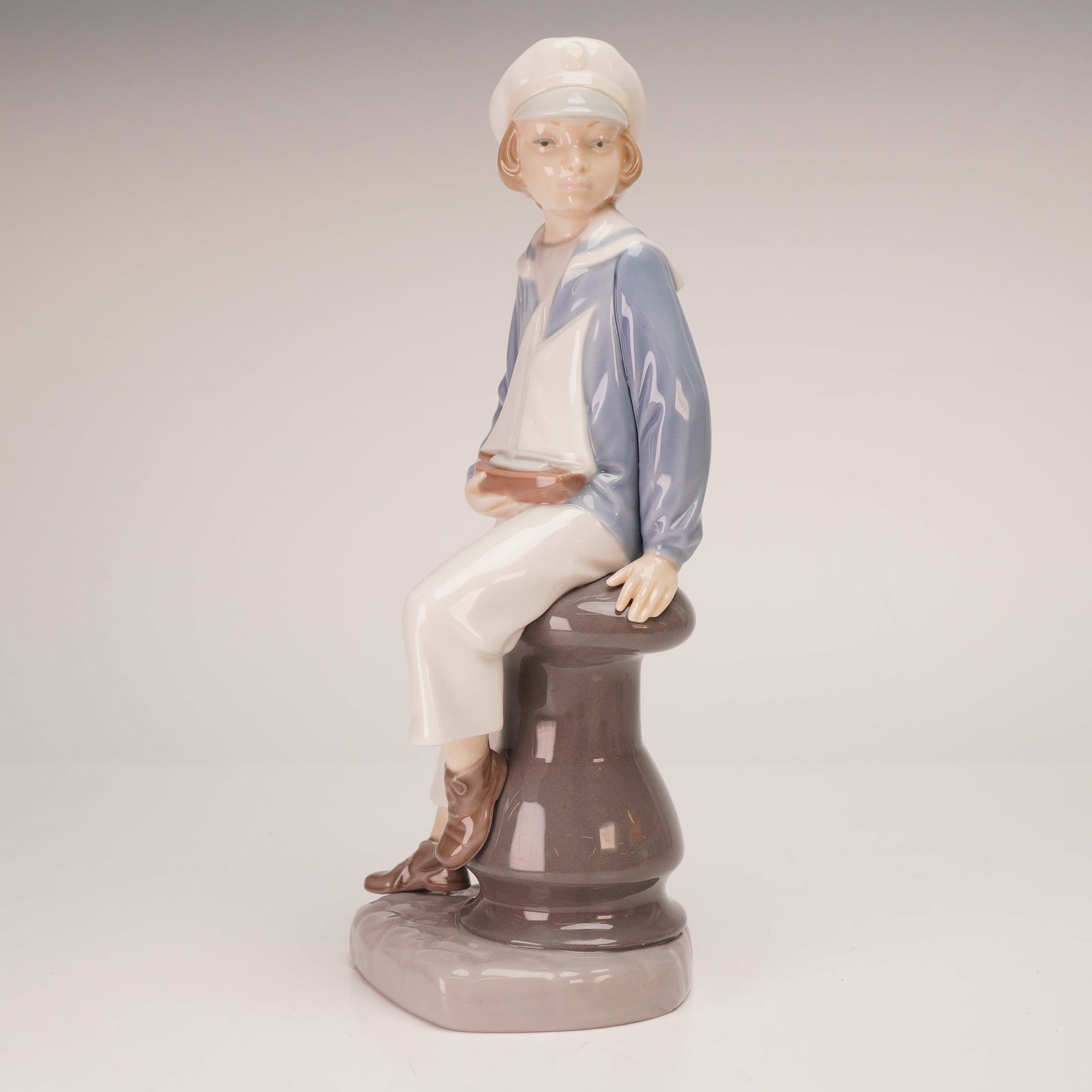 Lladro Porcelain Figurine, Boy with Yacht 1004810: A glazed porcelain figurine depicting a young boy in a traditional sailor uniform. He is seated upon a brown mooring bollard while holding a small toy sailboat. The underside features the blue undergl