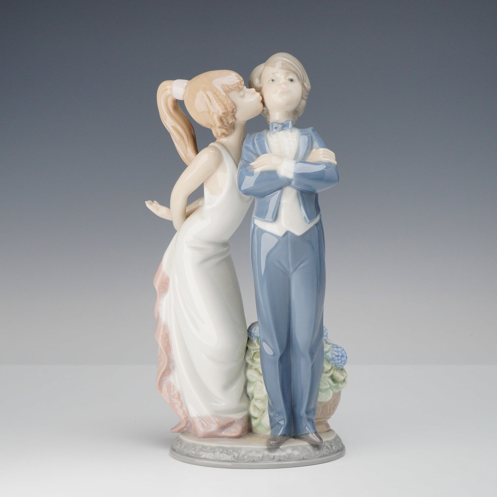 Lladro Porcelain Figurine, Let's Make Up 1005555: This Lladro glazed porcelain figurine, depicts a young boy in a formal blue suit and bowtie standing with arms crossed, while a girl in a cream-colored gown with a pink sash leans in to kiss his cheek