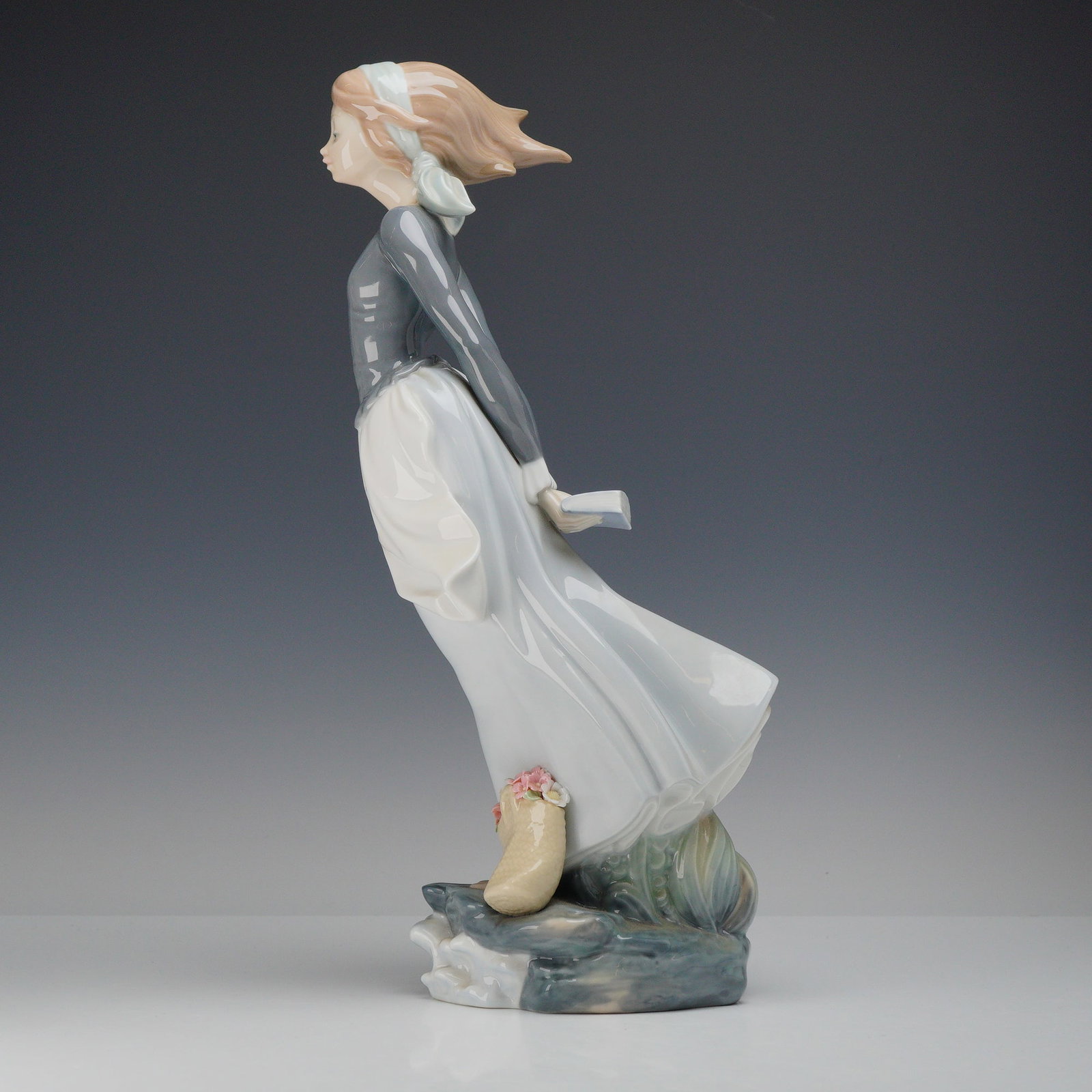 Lladro Porcelain Figurine, Sea Breeze 4922: The figurine depicts a young woman in a contemplative pose, standing atop a rocky, wave-lapped base. Her long, pale blue skirt and hair, bound by a white headband, are swept back as if by a strong bre