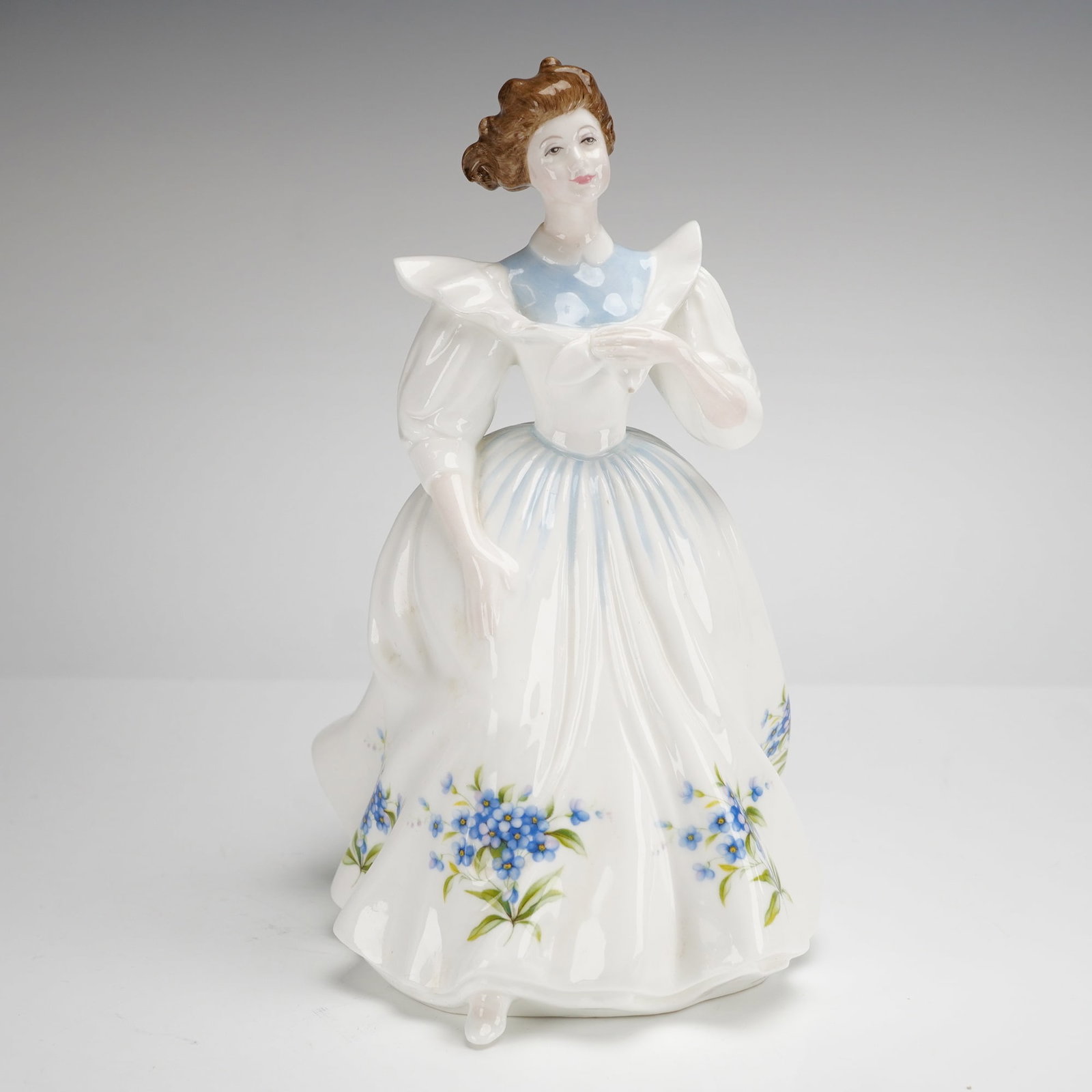 Royal Doulton Bone China Figurine July HN2794: This Royal Doulton bone china figurine, titled July (HN 2794) from the Figure of the Month series, was modeled by Peggy Davies and is dated 1988. The piece depicts a standing woman in a white gown fea